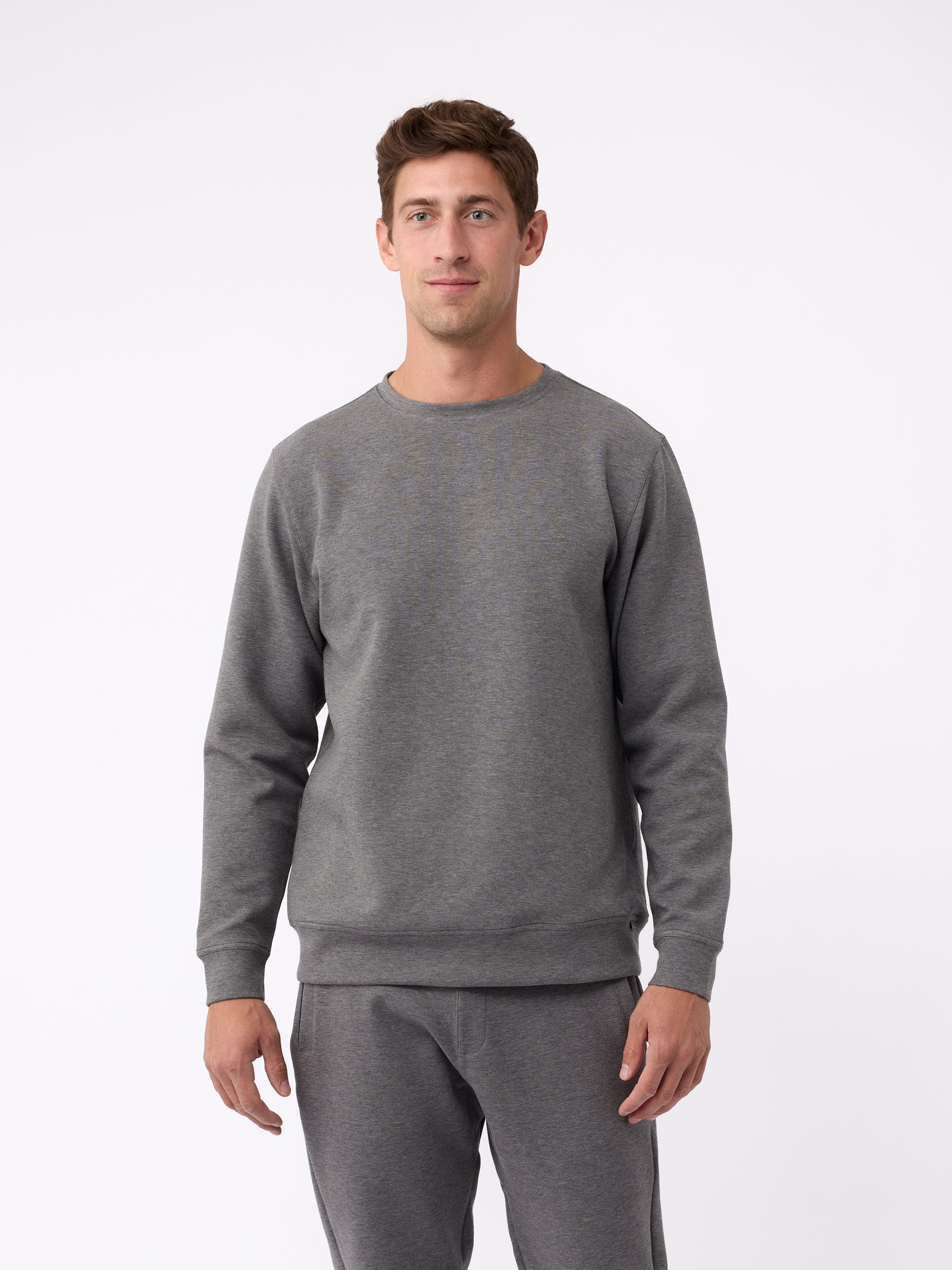 A man is wearing the Men's StretchTech Crewneck sweatshirt from Cozy Earth, paired with matching sweatpants. He stands against a plain white background with short brown hair, looking forward with a neutral expression. 