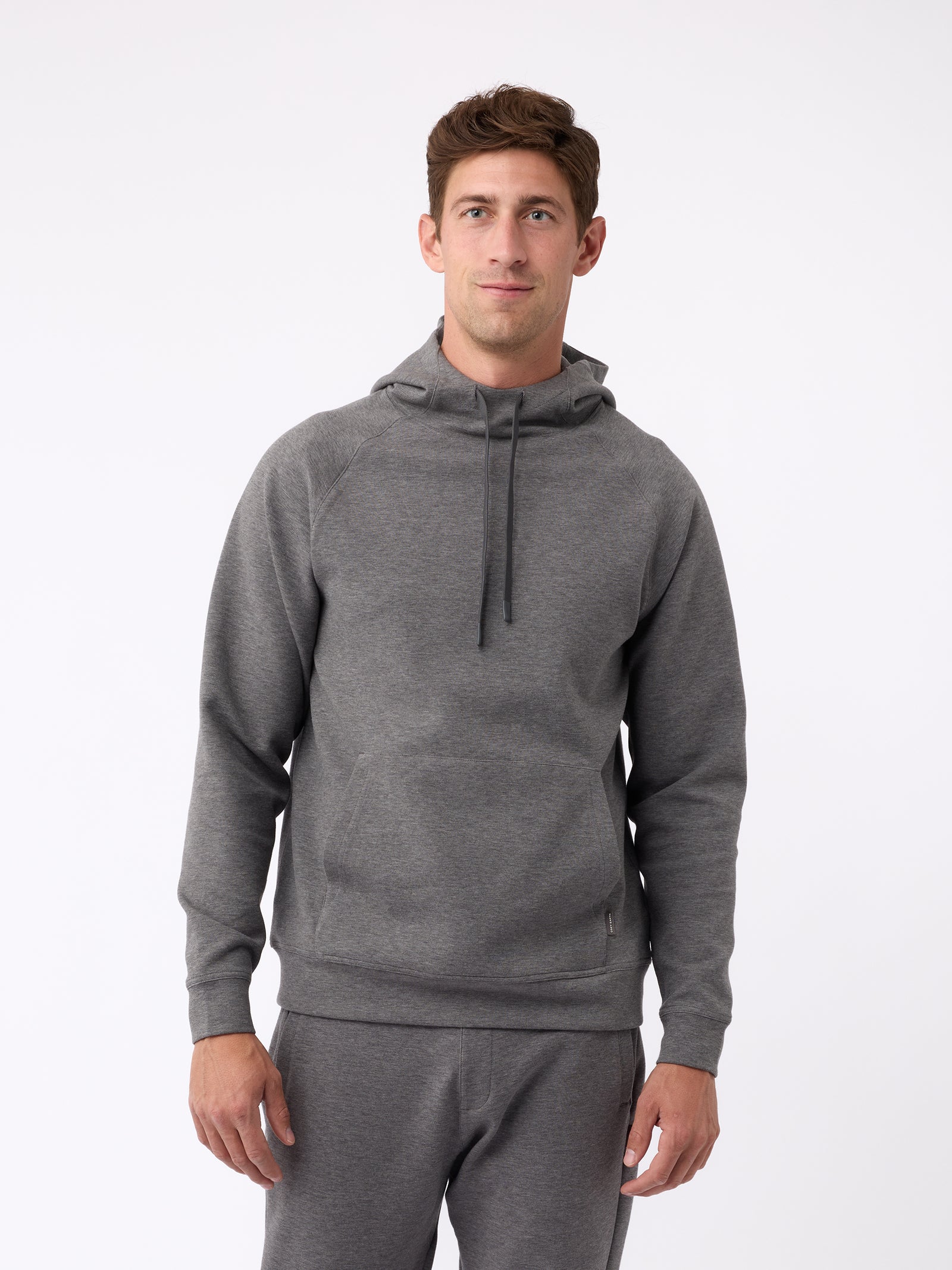 A person wearing the Men's StretchTech Hoodie from Cozy Earth, paired with matching gray pants, stands against a plain white background. The hoodie features drawstrings and a front pocket. The individual is looking forward with a neutral expression. 