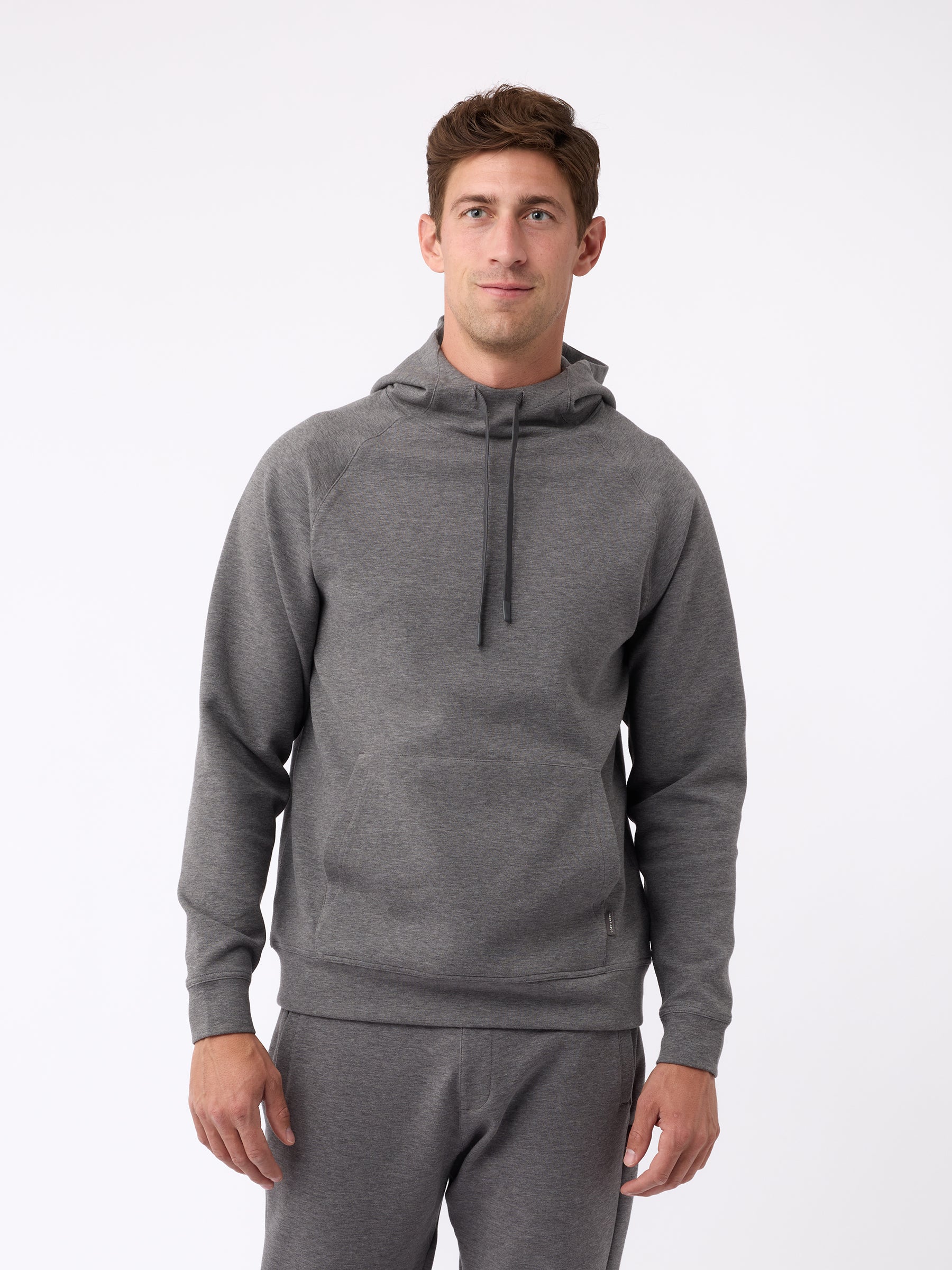 A person wearing the Men's StretchTech Hoodie from Cozy Earth, paired with matching gray pants, stands against a plain white background. The hoodie features drawstrings and a front pocket. The individual is looking forward with a neutral expression. |Color:Coal Heather