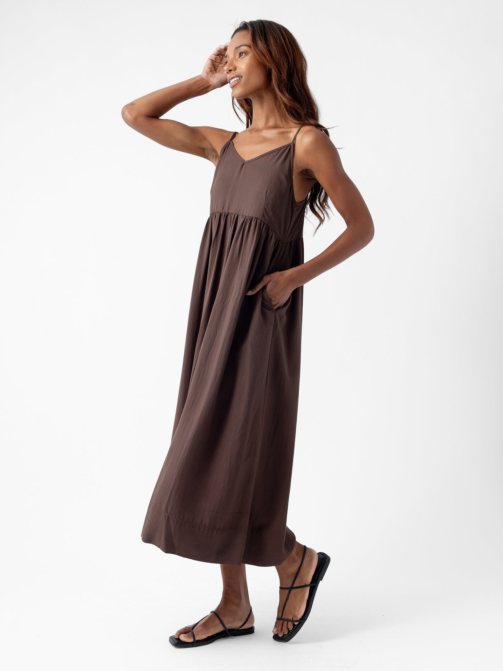 A person stands against a white background wearing the Women's Sunset Gathered Dress by Cozy Earth. The sleeveless, brown maxi dress features convenient pockets. They are smiling and looking off to the side with one hand raised to their head and the other resting on a pocket. They are wearing black sandals, and their long hair flows down. 