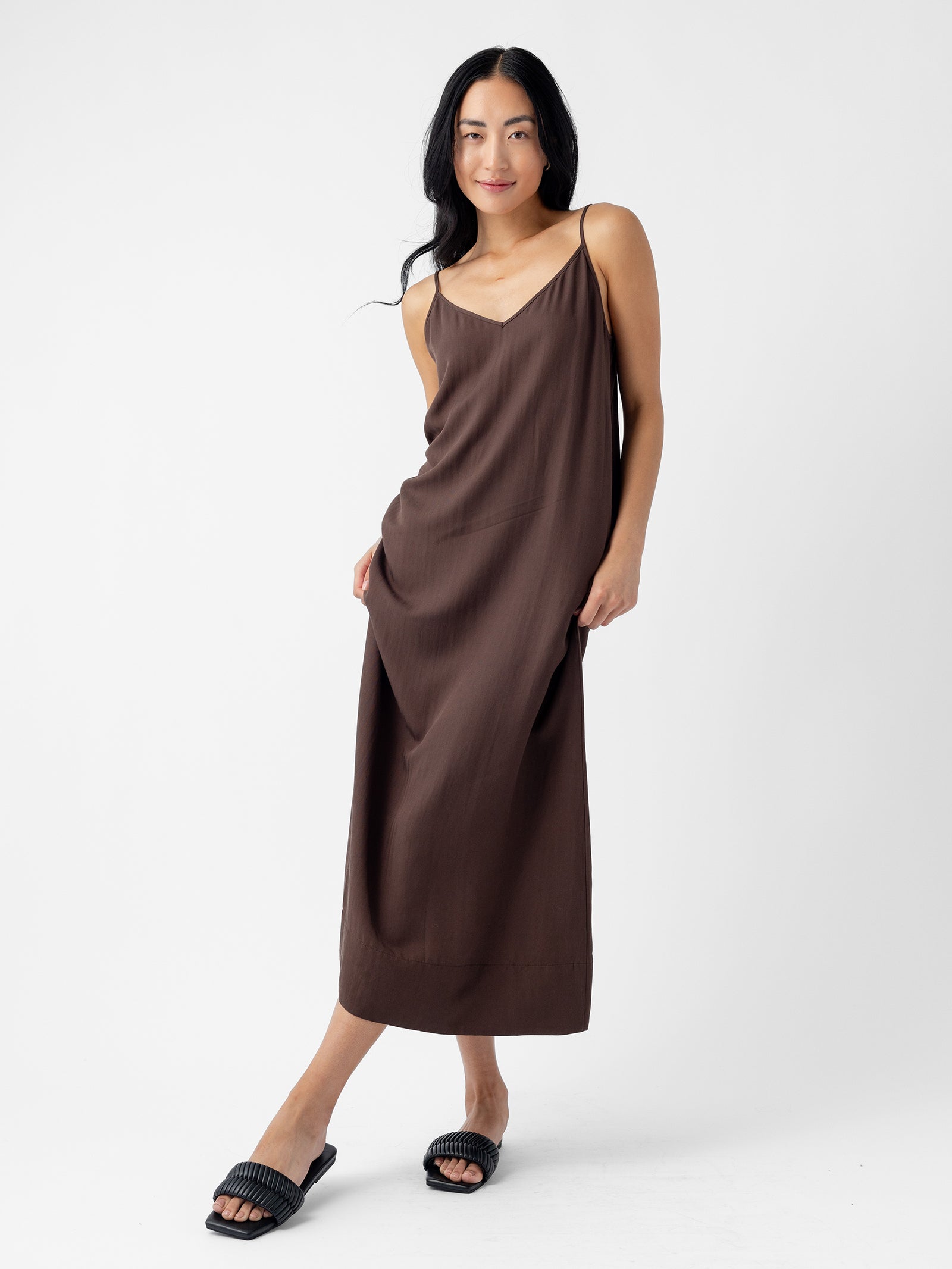 A woman with long, dark hair stands against a white background. She is wearing Cozy Earth's Women's Sunset Slip Dress in a loose, brown spaghetti strap design that reaches her ankles and black slide sandals. She is slightly leaning to one side, with one hand in her pocket and the other touching the dress. 