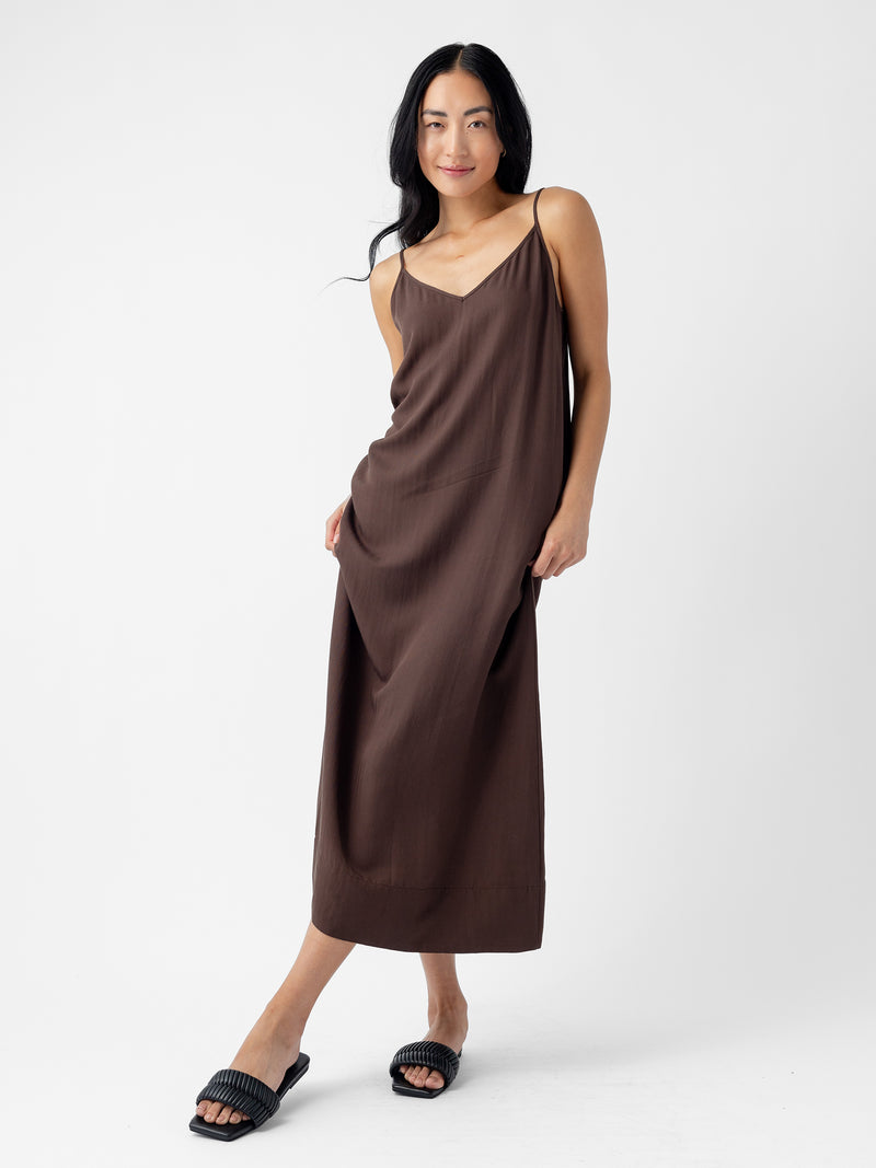 A woman with long, dark hair stands against a white background. She is wearing Cozy Earth's Women's Sunset Slip Dress in a loose, brown spaghetti strap design that reaches her ankles and black slide sandals. She is slightly leaning to one side, with one hand in her pocket and the other touching the dress.