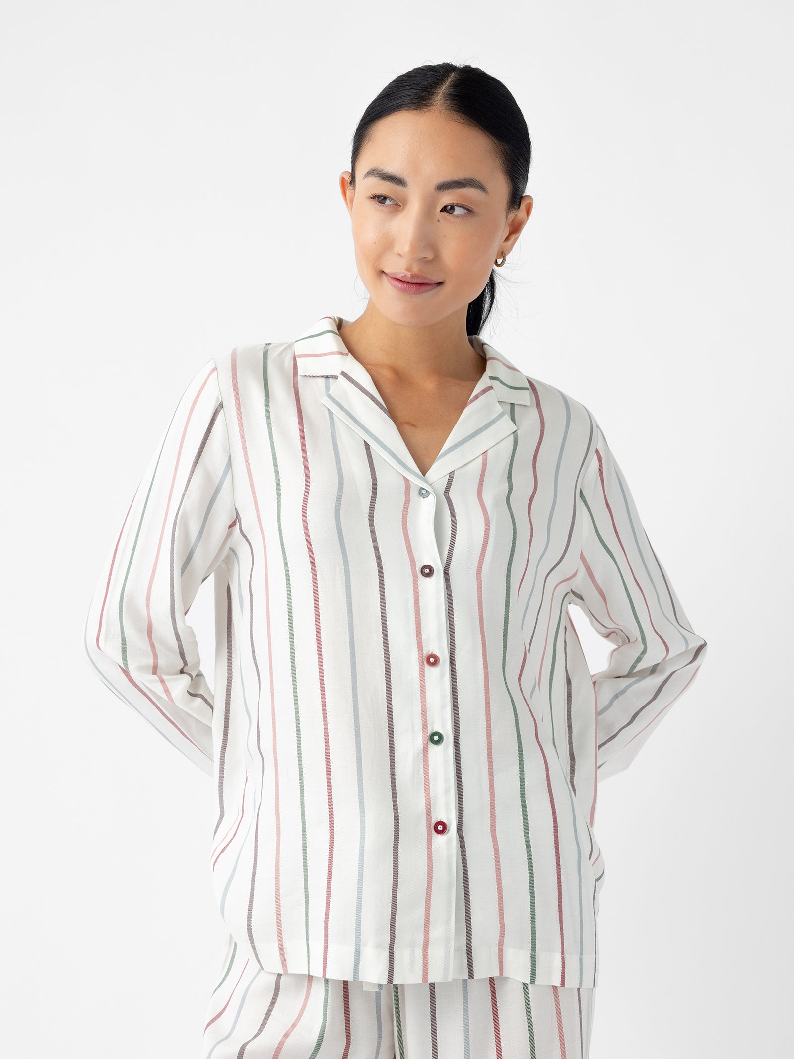 A person is wearing the Women's Soft Woven Long Sleeve Pajama Top by Cozy Earth, featuring vertical multicolored stripes on a white background with button detailing. The individual is looking to the side with their hands behind their back. 