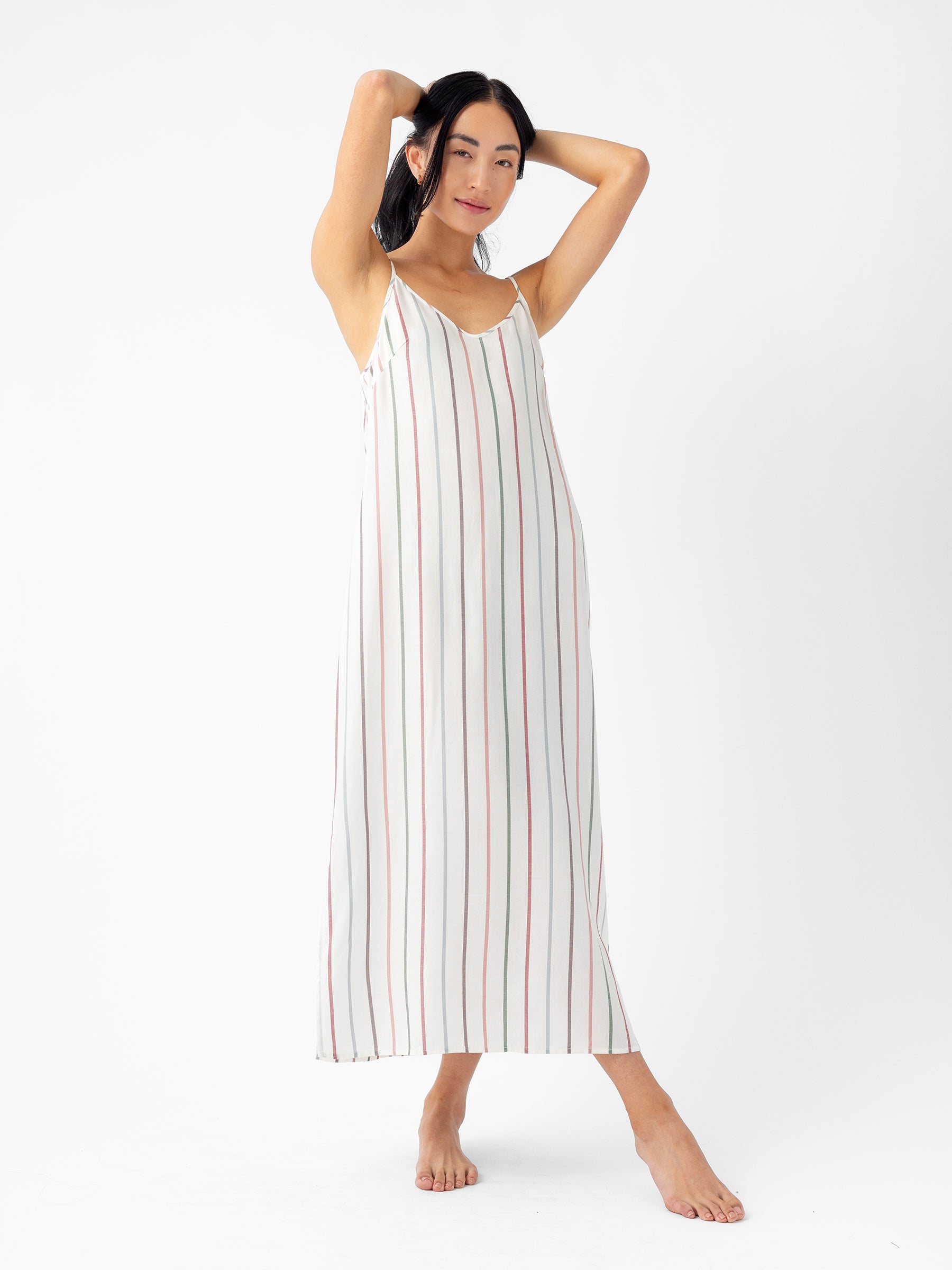 A person poses barefoot against a plain white background, with arms raised behind their head, wearing a long, sleeveless Women's Soft Woven Nightgown by Cozy Earth. The nightgown features vertical red, blue, and black stripes on a white base. |Color:Cottage Stripe