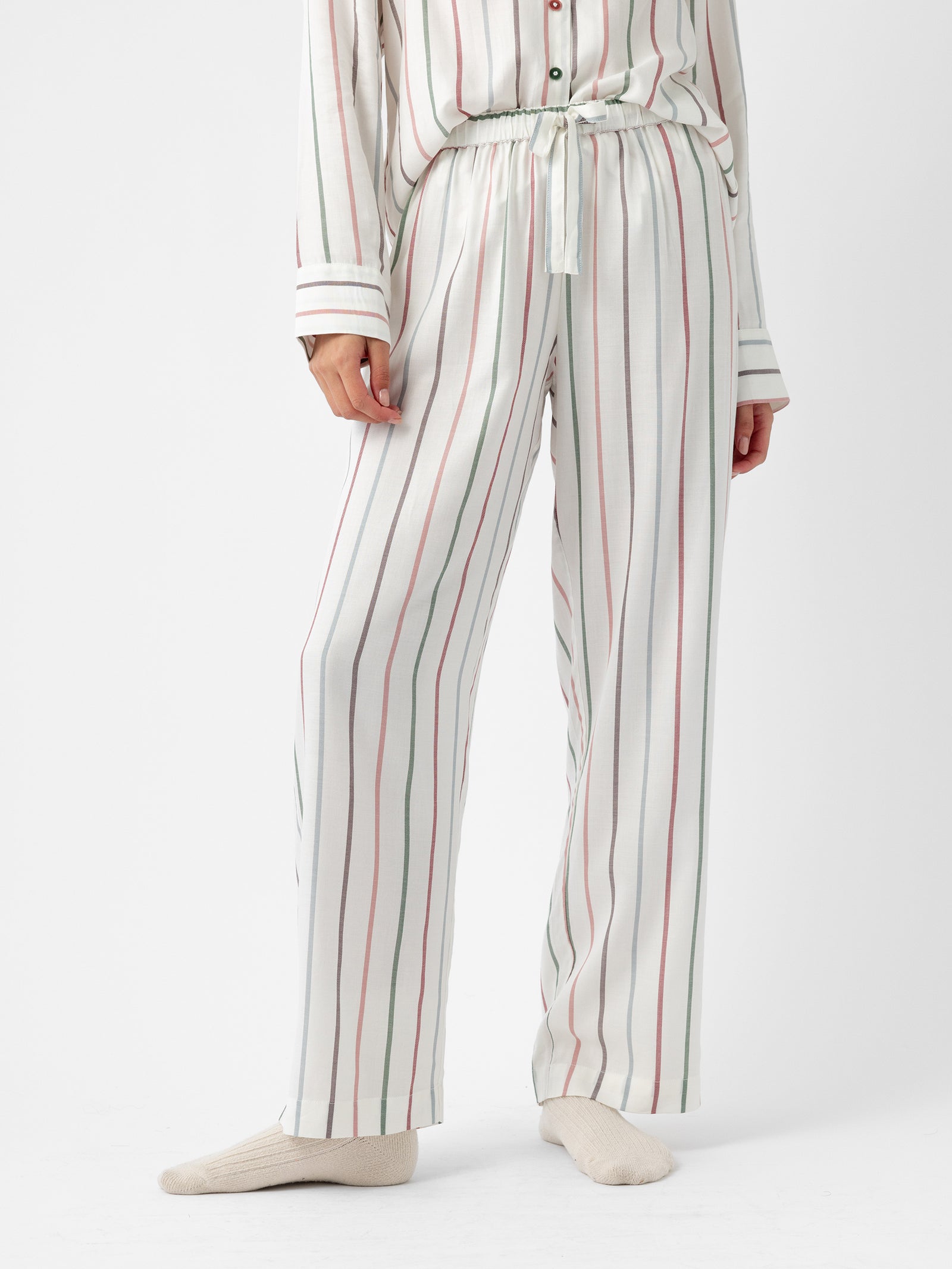 A person wearing Cozy Earth's Women's Soft Woven Pajama Pant, featuring a white fabric with vertical red and green stripes, paired with a matching long-sleeved top. They are standing against a plain white background and wearing light-colored socks. 