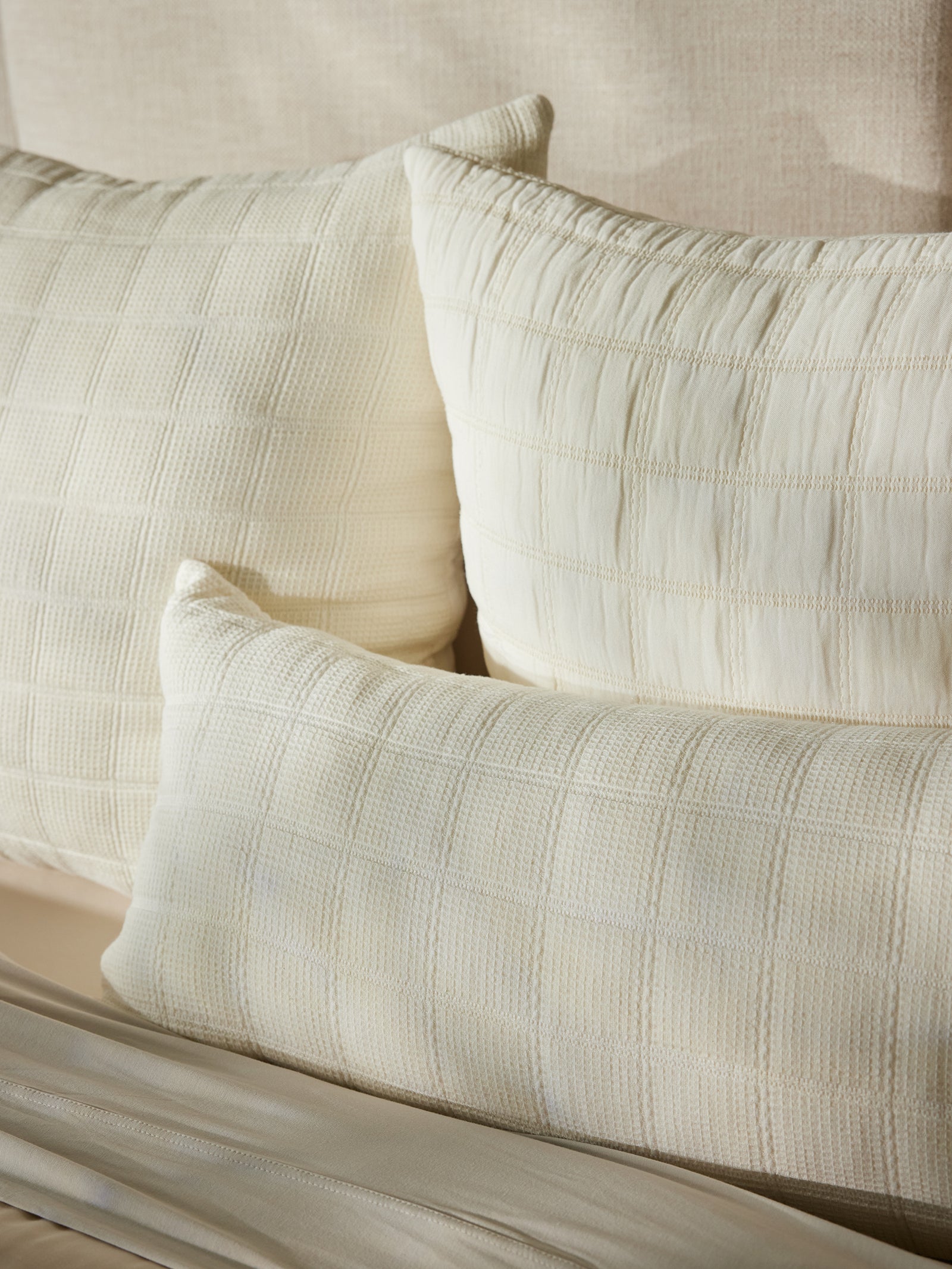 Three Cozy Earth Waffle Windowpane Pillows, textured and off-white, rest on a beige bed with sunlight casting soft shadows. The pillows feature a woven, checkered pattern and are arranged overlapping each other, creating a cozy and inviting atmosphere. 