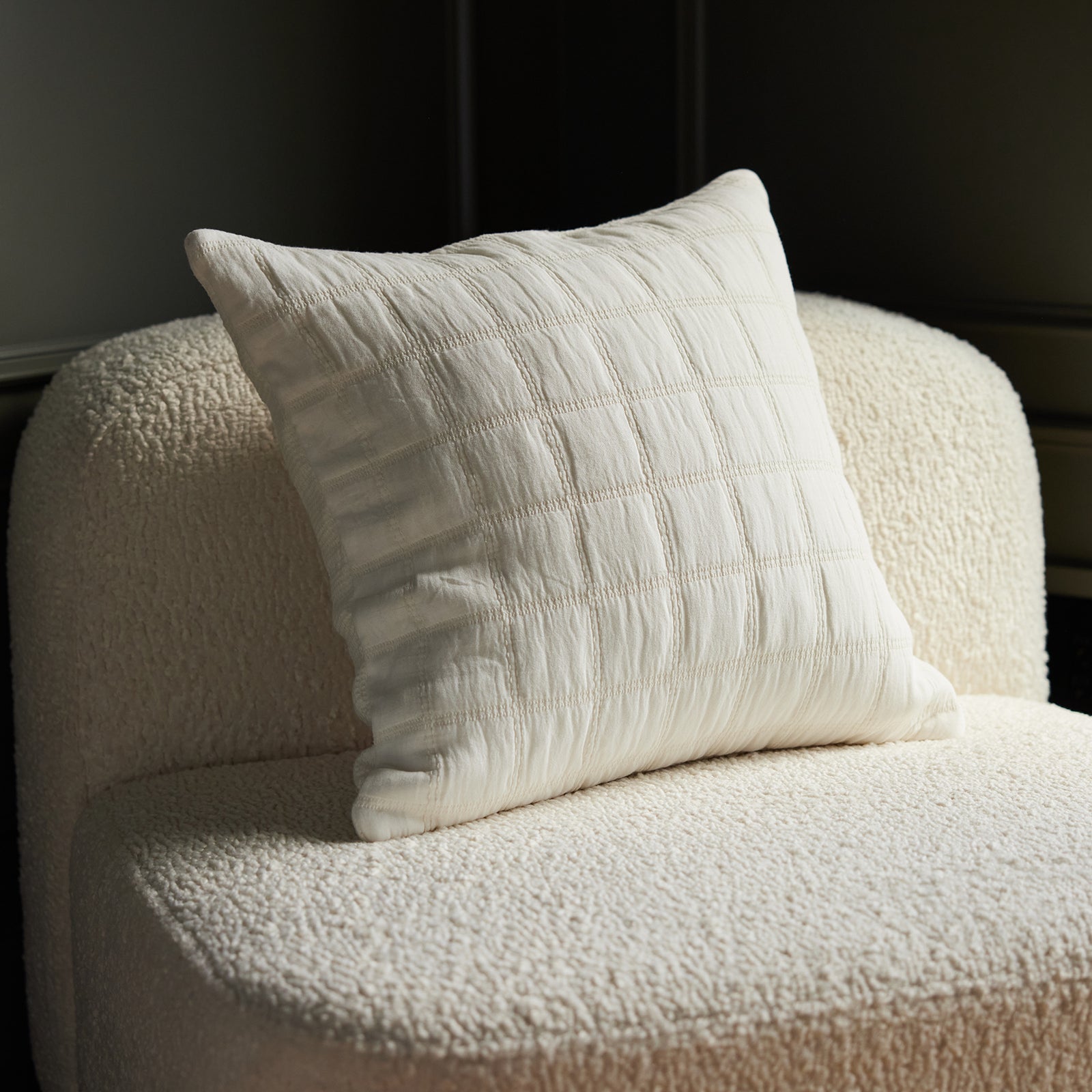 The Cozy Earth Waffle Windowpane Pillow, with its white textured grid pattern, is positioned on a cream, plush armchair in a softly lit room. Together, the pillow and chair create a cozy and inviting atmosphere. 