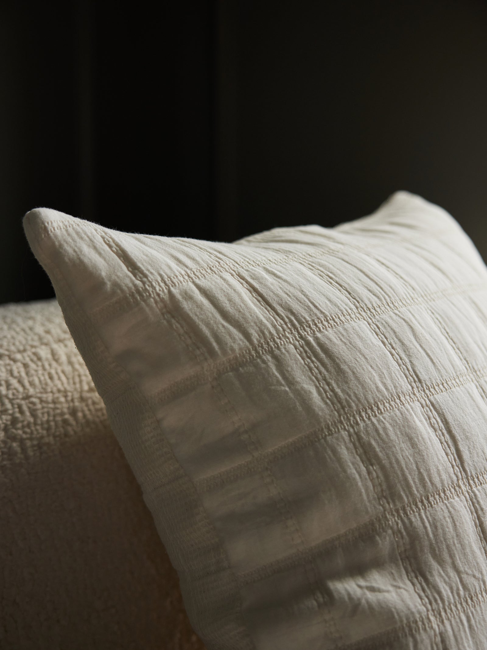 A close-up of the Cozy Earth Waffle Windowpane Pillow, showcasing its white cushioned design with a textured, checkered pattern. The pillow is placed on an off-white, textured surface, with dim lighting creating soft shadows. The dark background contributes to a cozy and warm ambiance. 