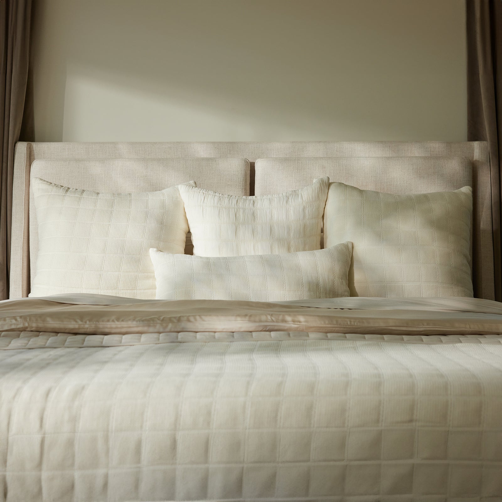 A neatly made bed with beige and white bedding, featuring four large pillows and a smaller Cozy Earth Waffle Windowpane Pillow. The bedspread and pillows showcase a subtle, textured grid pattern, while the soft natural light enhances the serene atmosphere. 