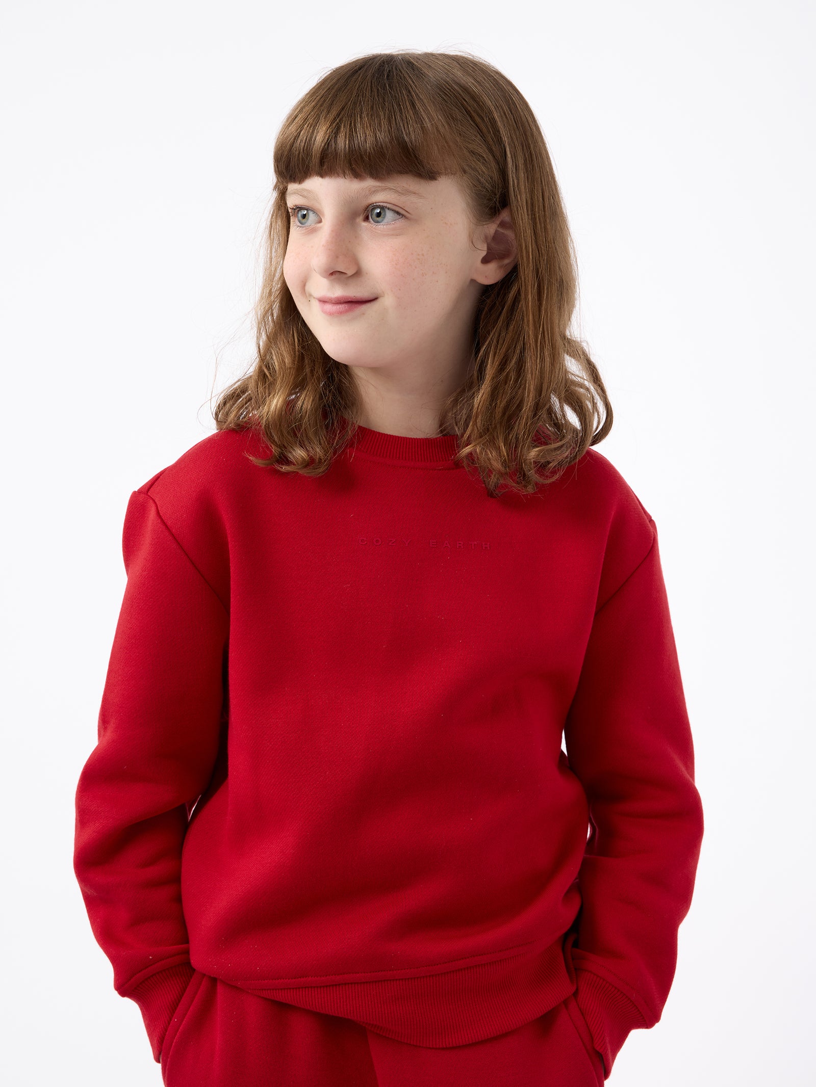 A young person with brown hair stands wearing a bright red Kid's CityScape Crewneck from Cozy Earth and pants against a plain white background, looking to the side and smiling slightly, with their hands tucked into their pockets. 