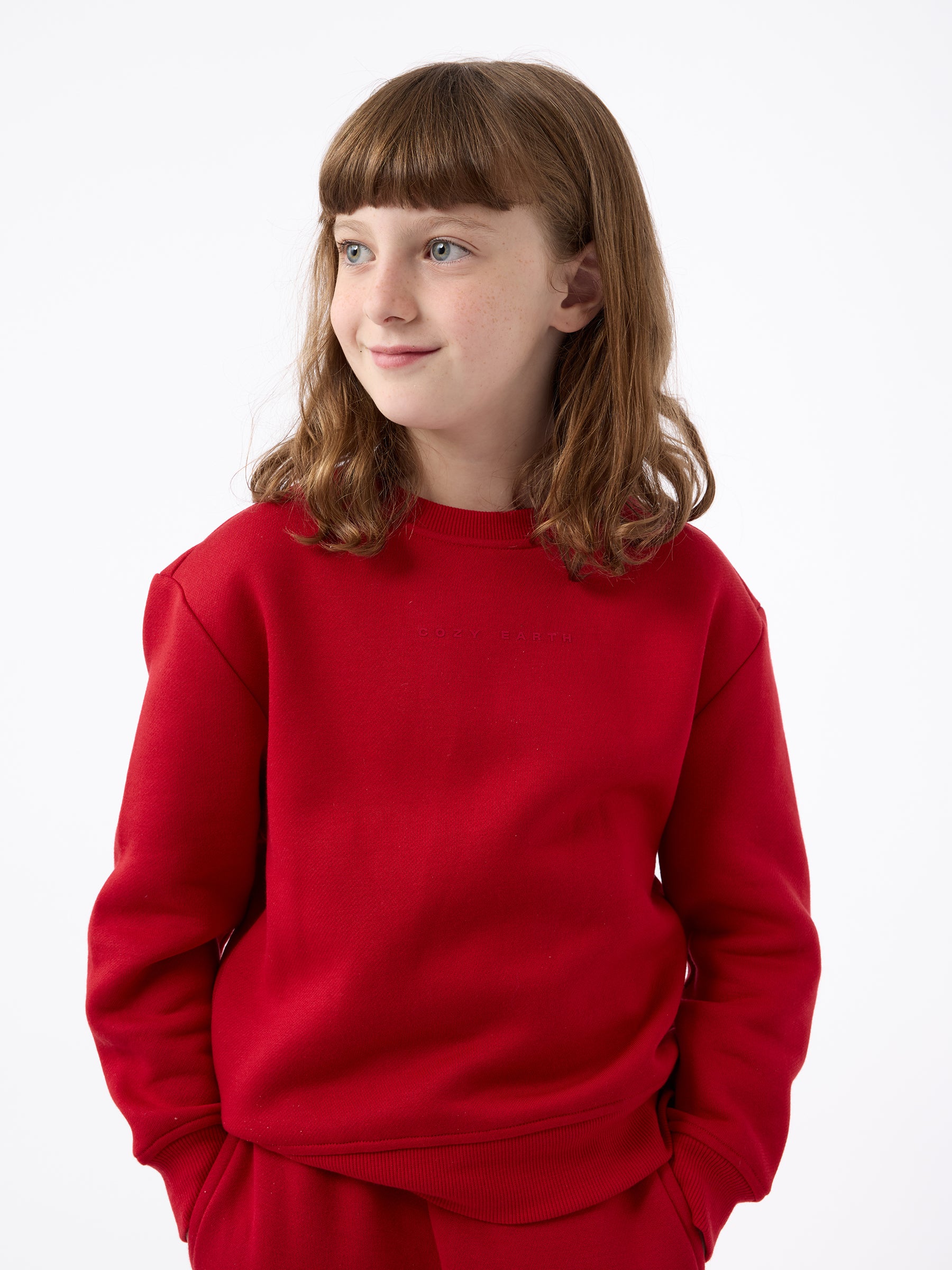 A young person with brown hair stands wearing a bright red Kid's CityScape Crewneck from Cozy Earth and pants against a plain white background, looking to the side and smiling slightly, with their hands tucked into their pockets. |Color:Crimson