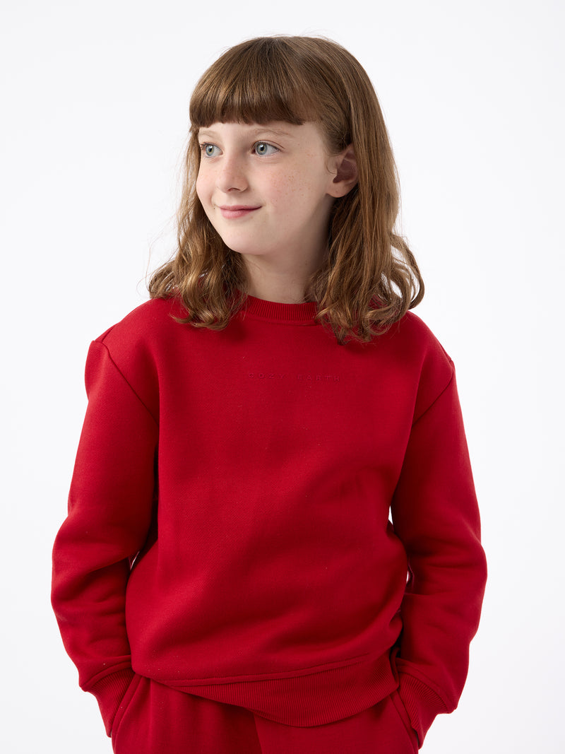 A young person with brown hair stands wearing a bright red Kid's CityScape Crewneck from Cozy Earth and pants against a plain white background, looking to the side and smiling slightly, with their hands tucked into their pockets.