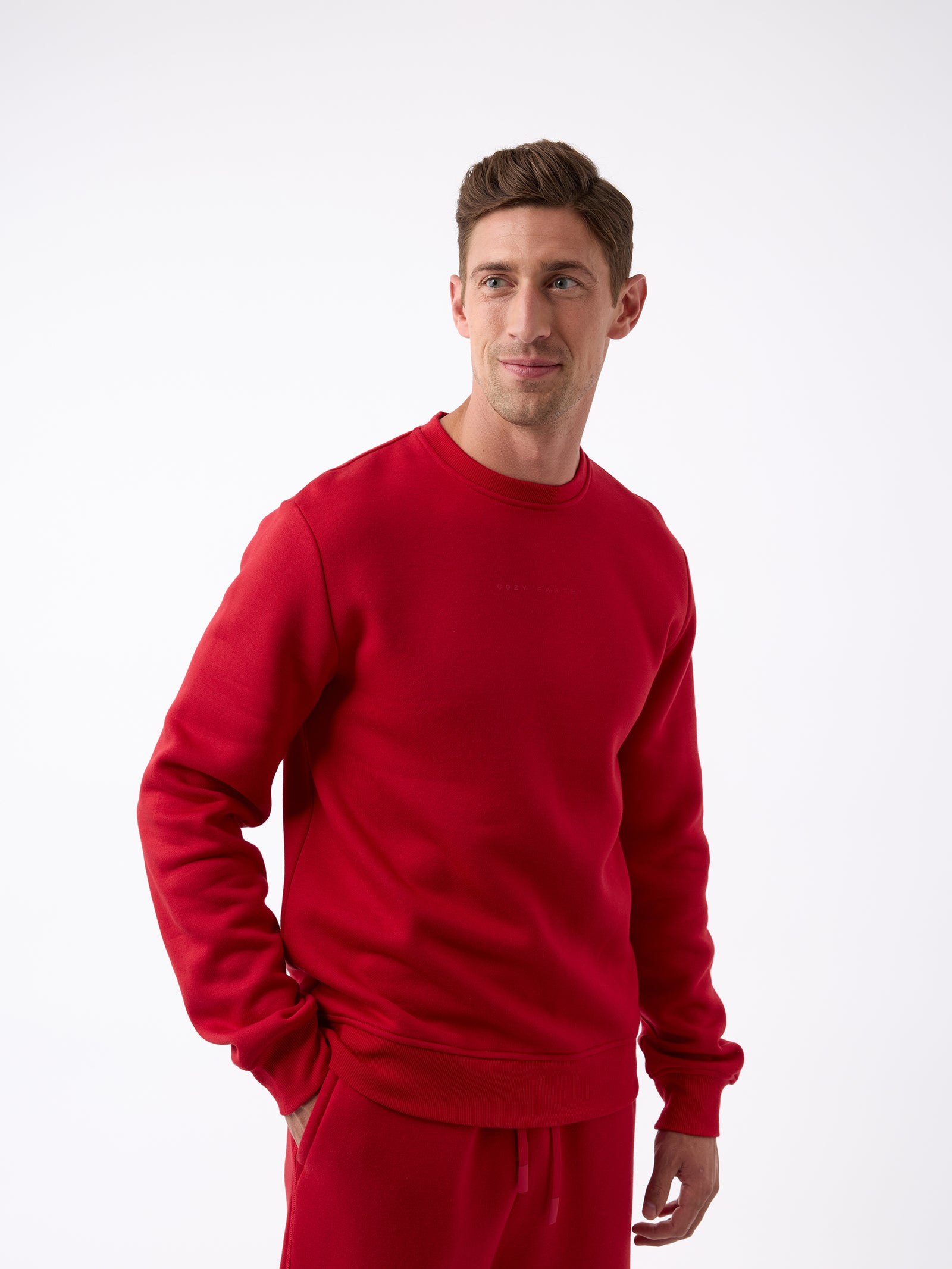 A person wearing a matching red Men's CityScape Crewneck and pants from Cozy Earth stands against a plain white background. They have short hair, are looking to the side with a slight smile, and have one hand in their pocket. 