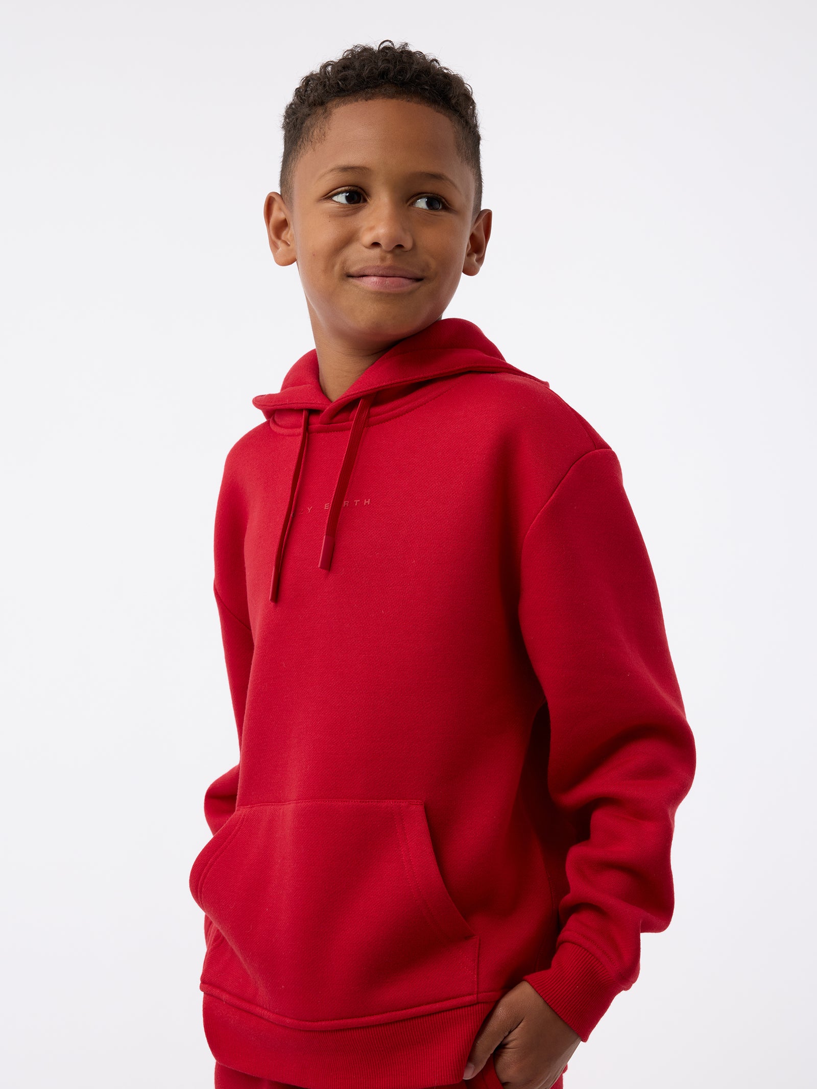 A child wearing a Kid's CityScape Hoodie by Cozy Earth smiles while looking to the side. The red hoodie features a front pocket against a plain white background. 