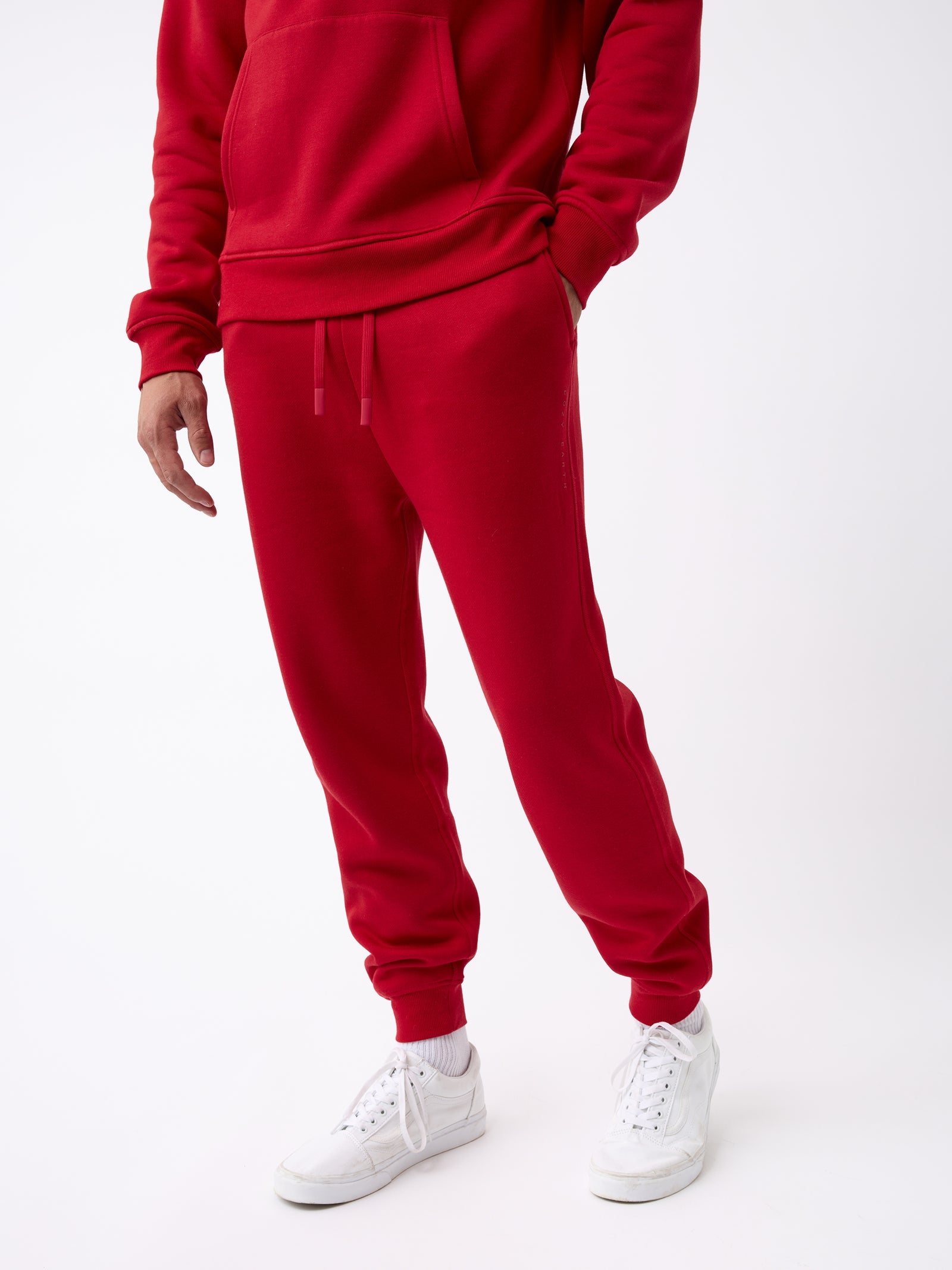 A person dressed in a Cozy Earth Men's CityScape Jogger set, featuring a red hoodie and matching red sweatpants, with hands casually tucked in the pockets. The ensemble is complemented by white socks and sneakers, standing against a plain light-colored background. 