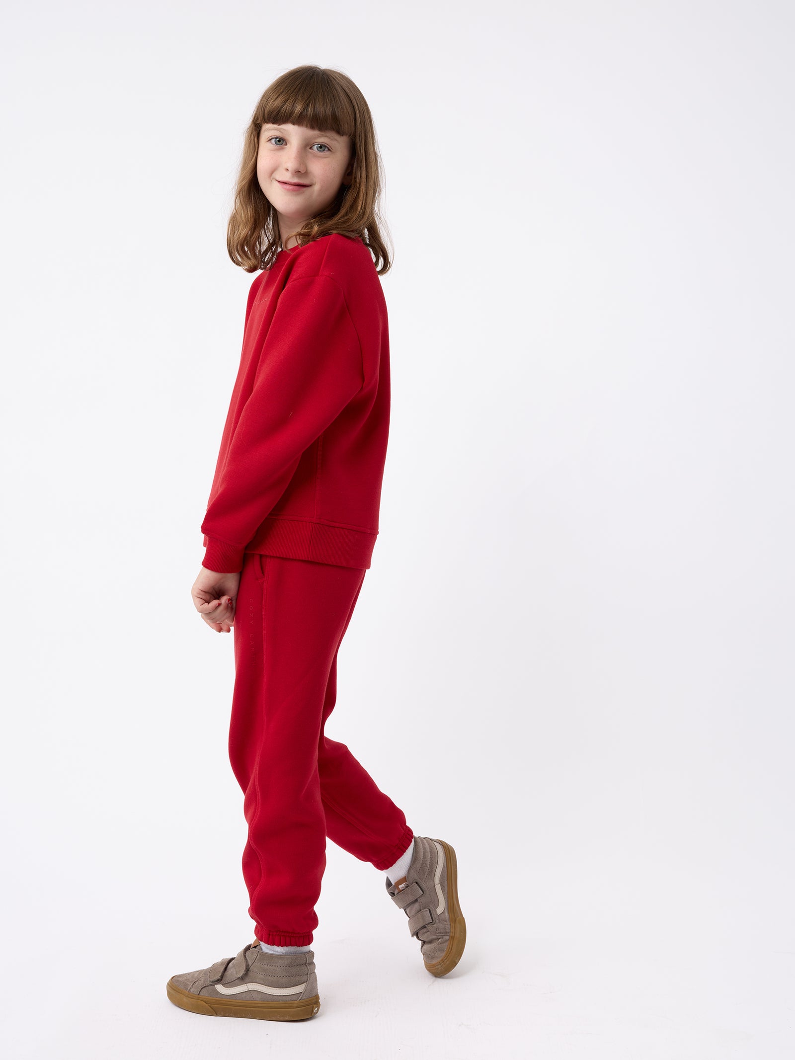 A child dressed in a vibrant red Cozy Earth CityScape Sweatpant outfit stands against a plain white background. They have shoulder-length brown hair and are looking towards the camera with a slight smile, complemented by their stylish brown sneakers. 