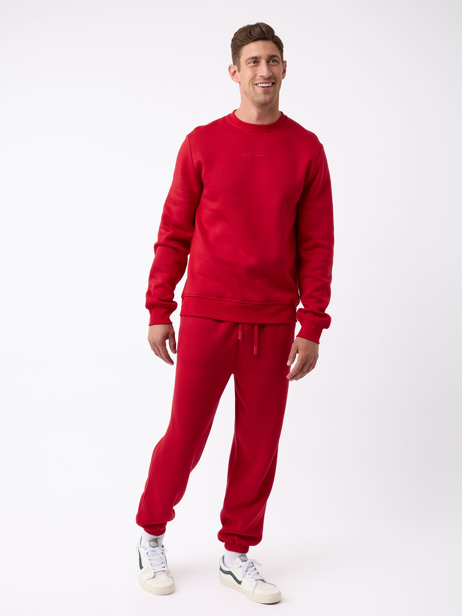 A person dressed in a matching red sweatshirt and Cozy Earth's Men's CityScape Sweatpant poses against a plain white background. They complement their outfit with white sneakers accented in green, smiling as they gaze slightly to the side. 