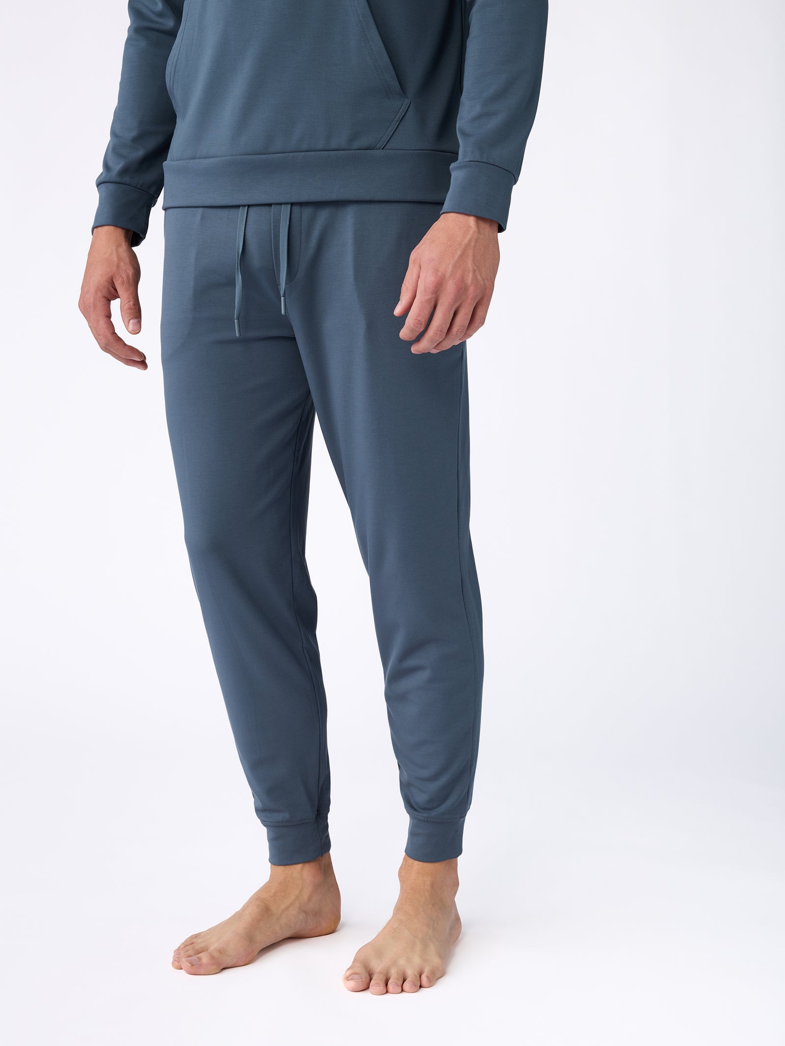 A person is wearing a matching Men's Ultra-Soft Bamboo Pullover Crew lounge set by Cozy Earth, featuring a long-sleeve top and jogger pants, standing barefoot against a white background. The top has a subtle texture and the pants include drawstrings, with the focus on the outfit from the neck down. 