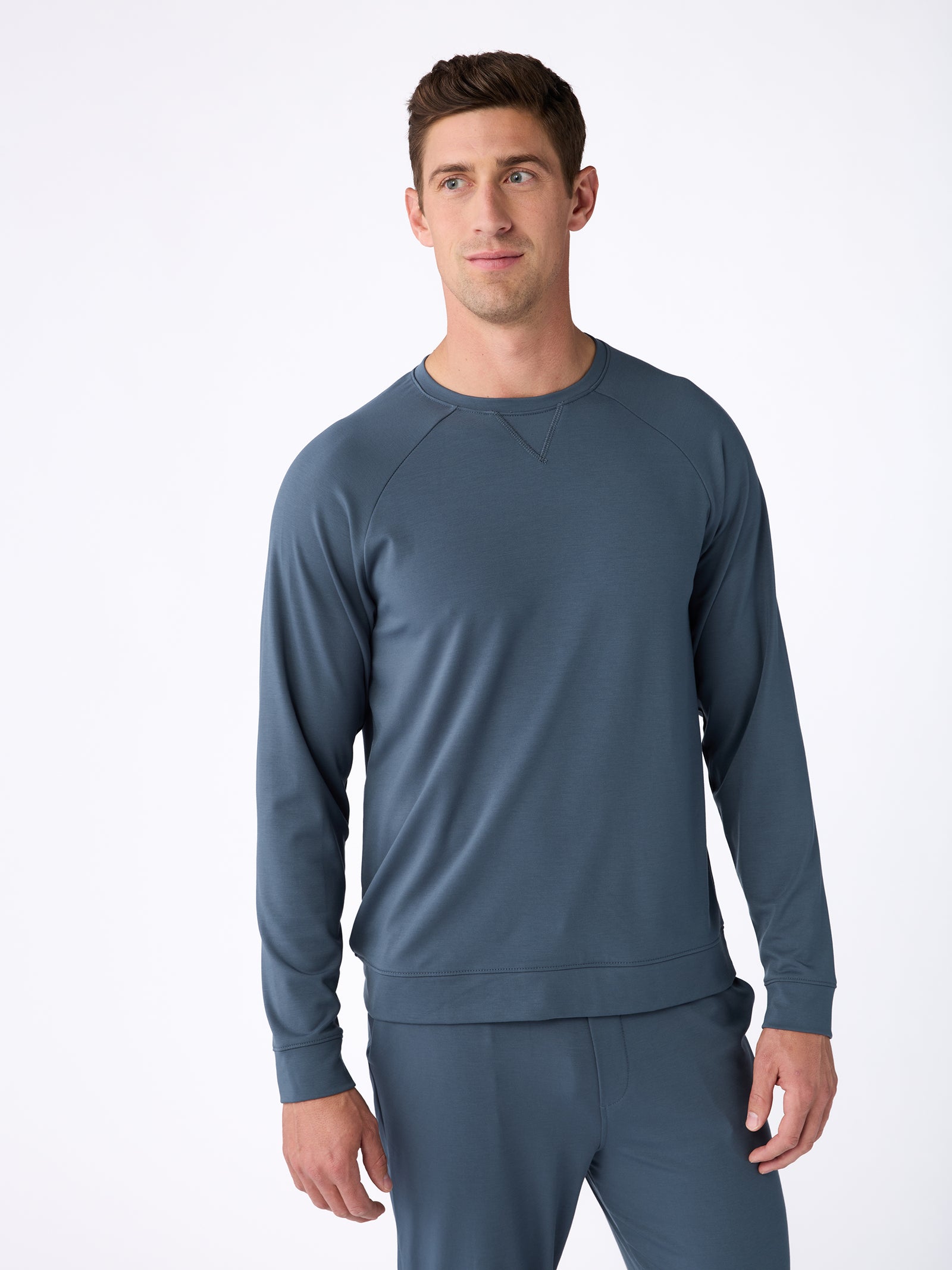 A man wearing a Men's Ultra-Soft Bamboo Pullover Crew from Cozy Earth in blue-gray, along with matching pants, stands against a plain white background. 