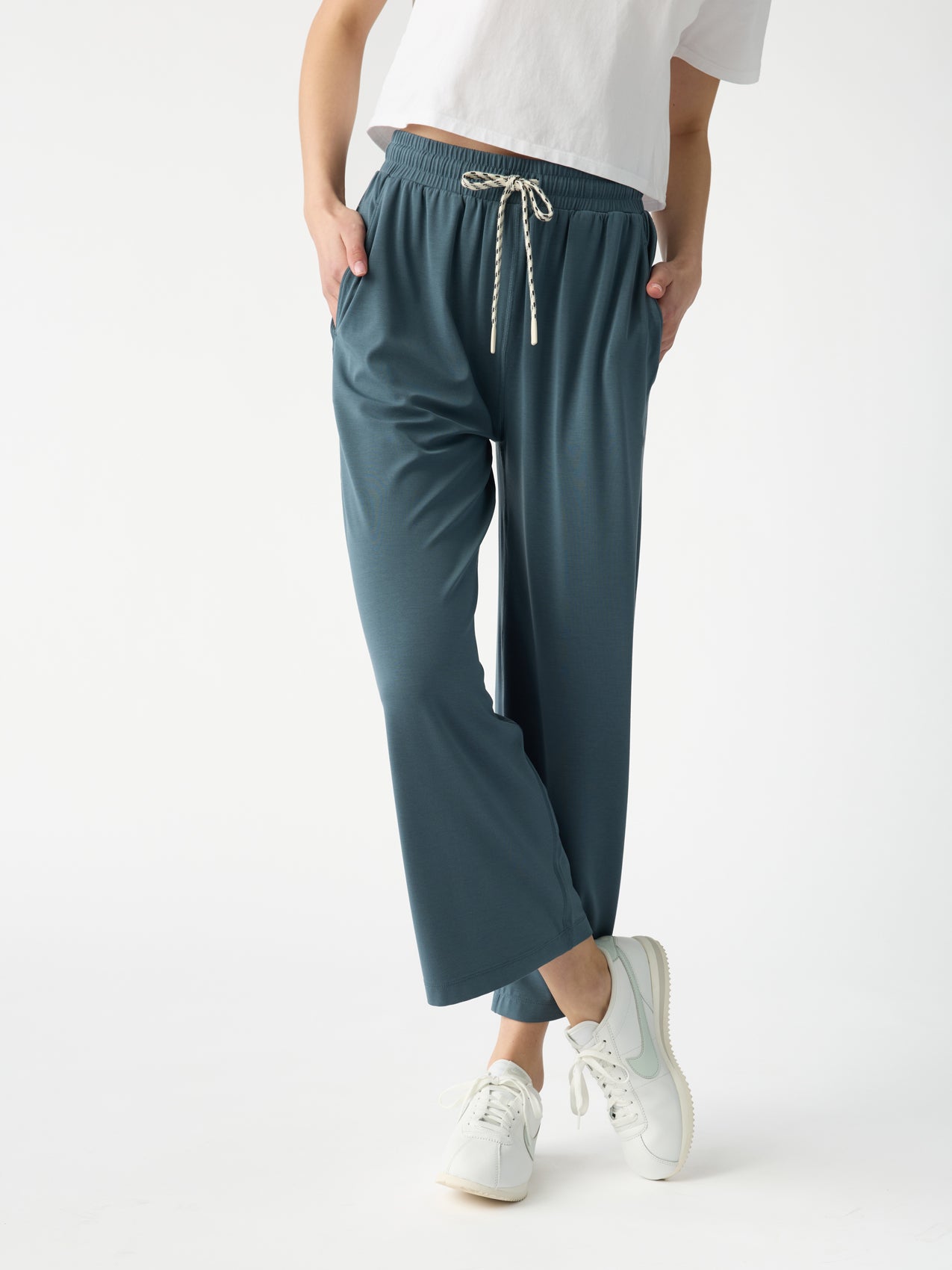 A person models the Cozy Earth Women's Studio Cropped Wide Leg Pant in teal, paired with a white T-shirt and sneakers. Their hands rest in their pockets against a plain white backdrop. |Color:Dawn