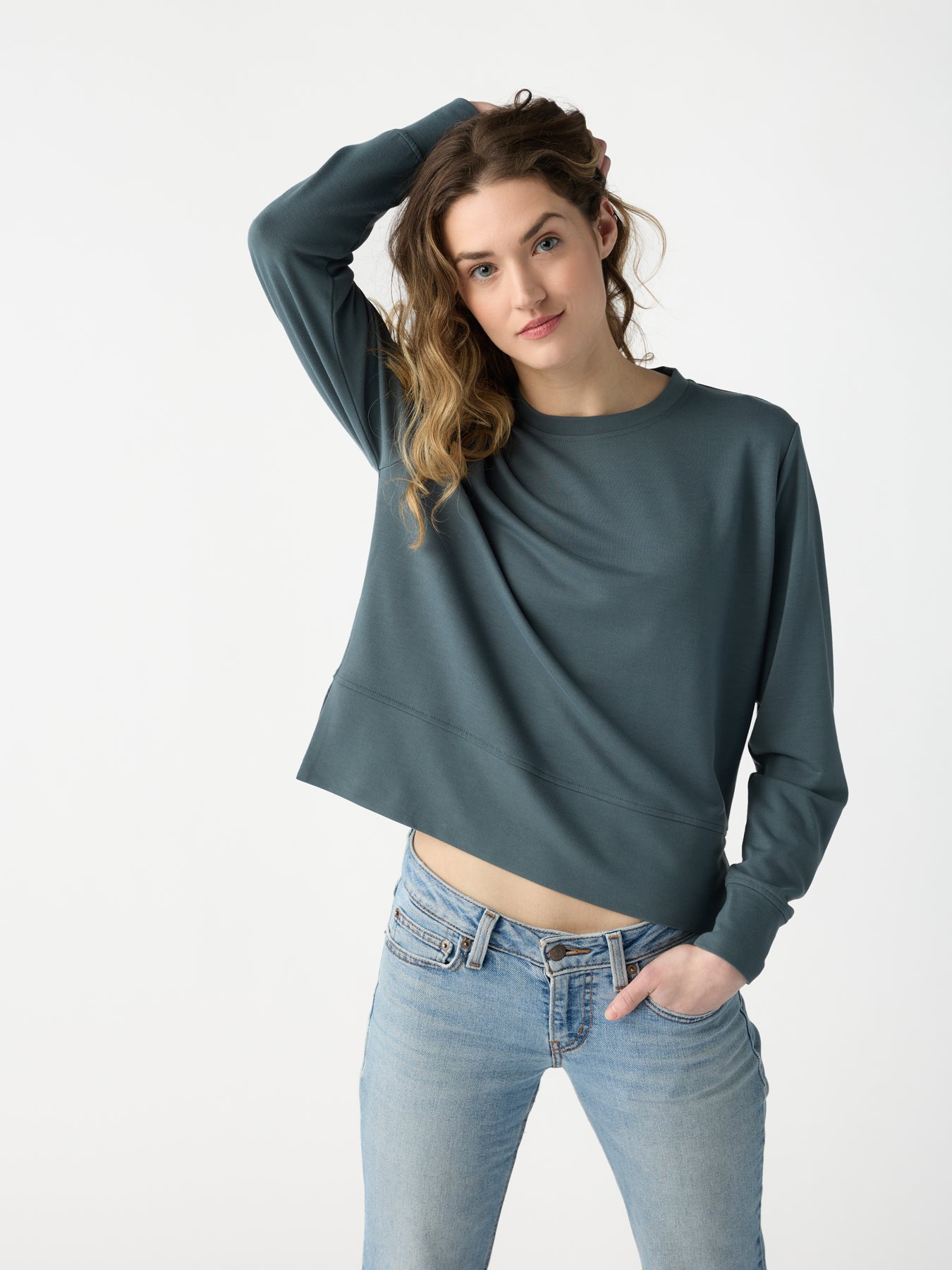 A person with wavy hair stands against a plain background, wearing a Cozy Earth's Women's StudioLite Crewneck in teal and light blue jeans. One hand rests behind their head while the other hangs relaxed at their side. They have a neutral expression. |Color:Dawn