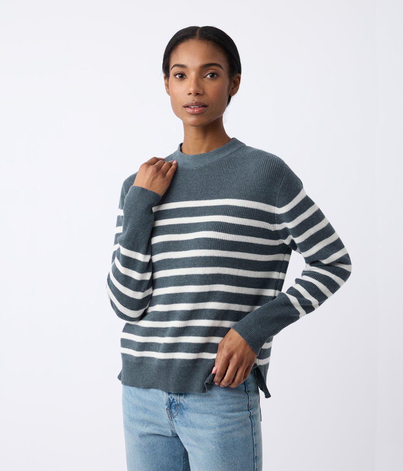 A person with long hair is wearing the Women's Rowan Sweater by Cozy Earth, featuring dark gray and white stripes, paired with light blue jeans. They stand against a plain white background, one hand on their neck and the other on their hip. |Color:Dawn/Bone Stripe