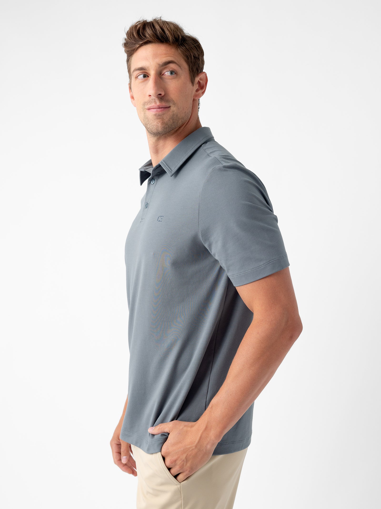 A man with short brown hair, dressed in a light gray Men's Everyday Polo from Cozy Earth and beige pants, poses against a plain white background. He looks slightly to his left, with one hand in his pocket. 