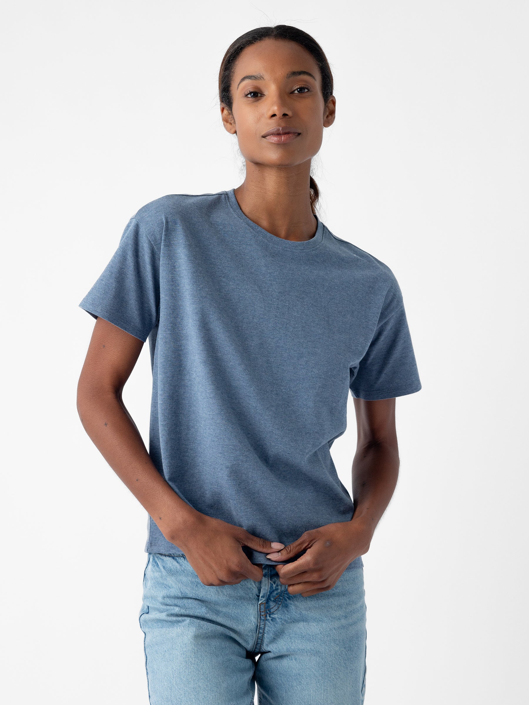 A person with long hair pulled back is wearing Cozy Earth's Women's All Day Tee, a blue short-sleeve t-shirt, along with light blue jeans. They are standing against a plain white background with a neutral expression, holding their hands near the waistband of their jeans. |Color:Deep Blue Heather