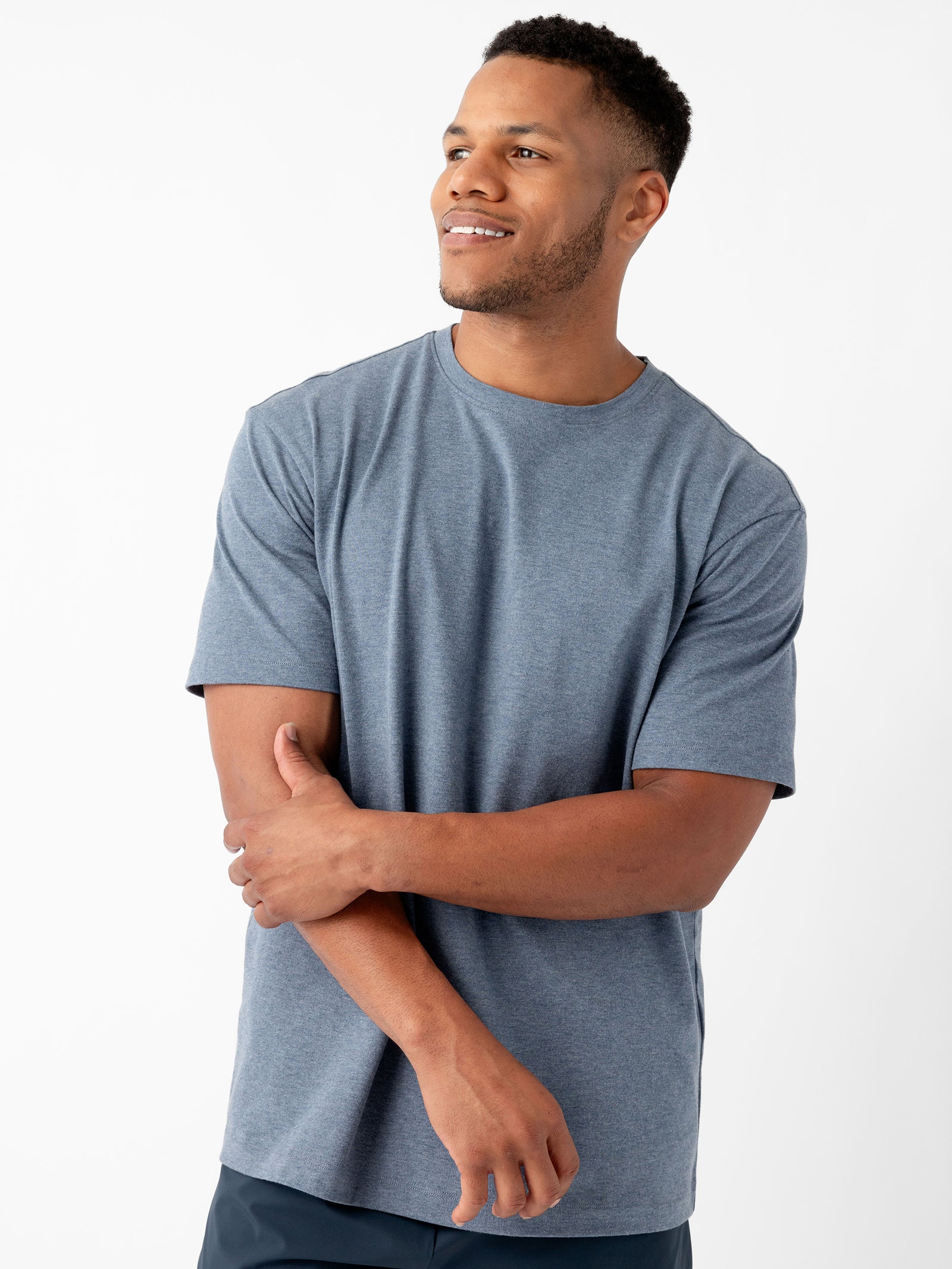 A man wearing the Cozy Earth Men's All Day Tee in plain blue-gray poses against a white background. He has short, dark hair and a beard, and he is smiling while looking off to the side. His left arm is bent, with his hand touching his right arm near the elbow. 