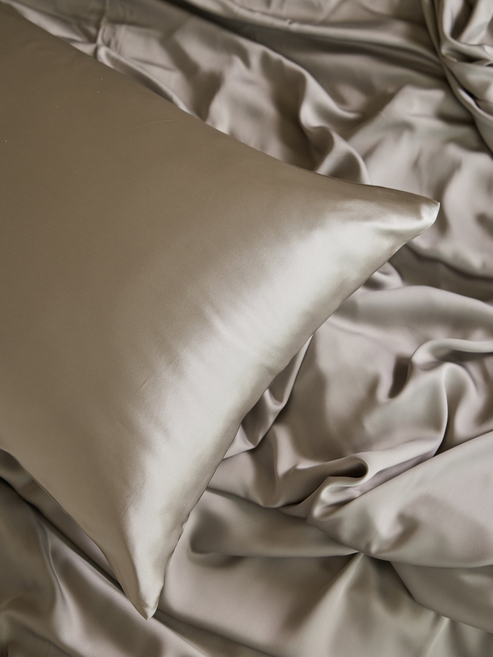 A smooth, silver Silk Pillowcase from Cozy Earth rests on matching sheets, creating a luxurious and elegant bed arrangement. The shiny texture of the fabric reflects light, highlighting its soft and sleek appearance. 