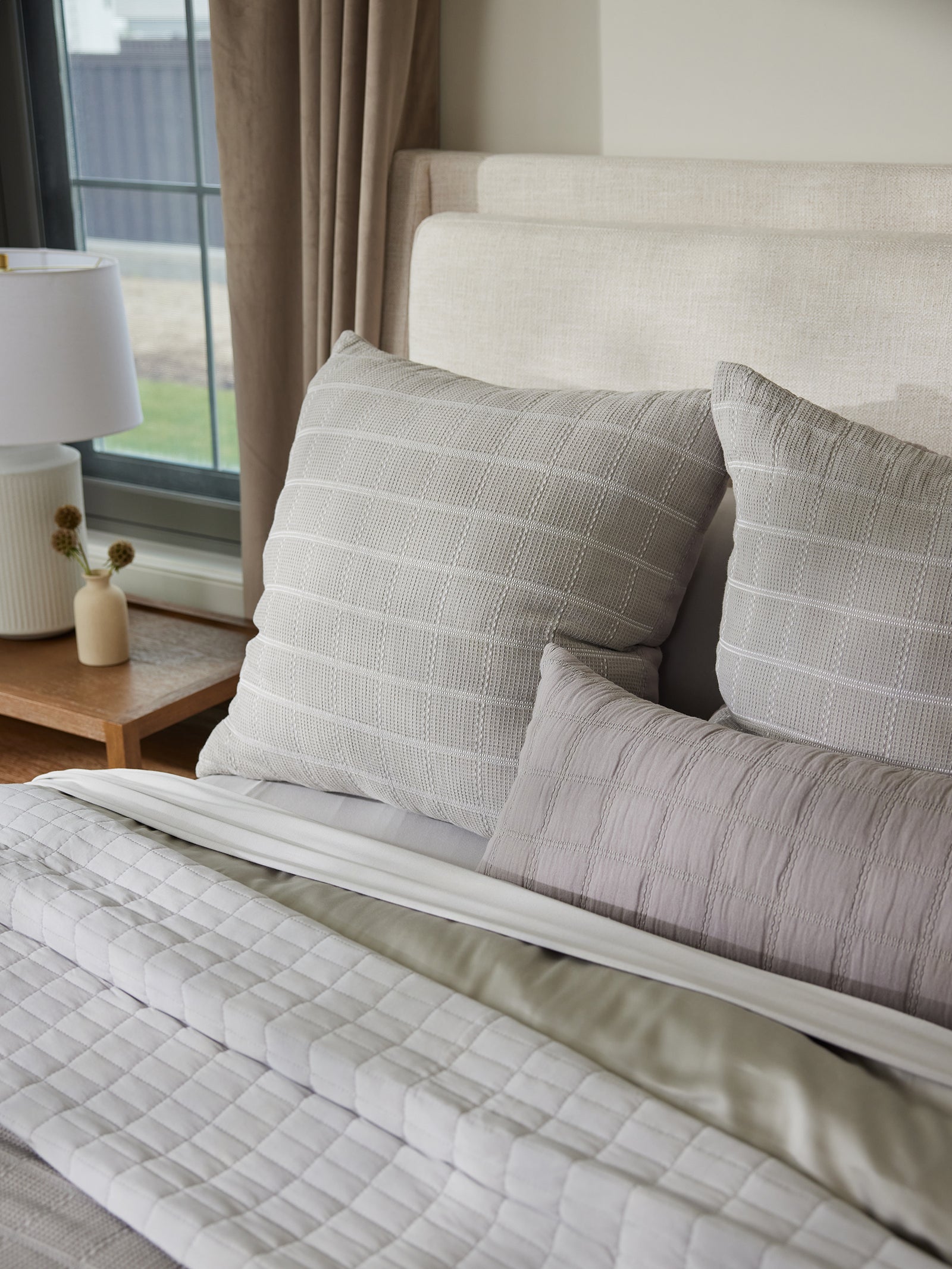 A neatly made bed with a white and gray color scheme, featuring three large Cozy Earth Waffle Windowpane Pillows and one smaller pillow. The bed is adorned with a white and gray quilt and positioned next to a wooden bedside table that holds a white lamp and a small plant, with a window in the background. 