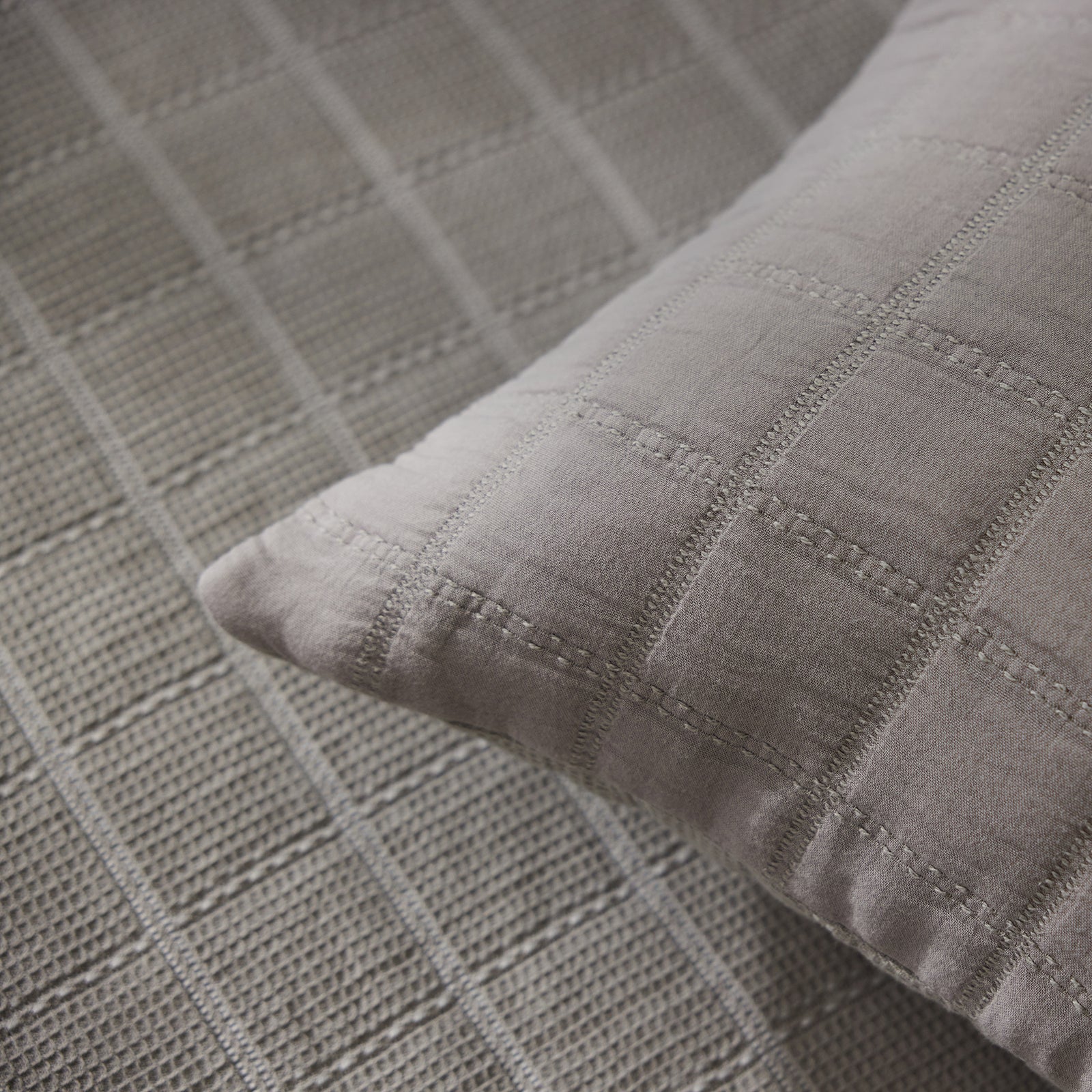 Close-up of the Cozy Earth Waffle Windowpane Pillow, featuring a grey grid pattern, placed on a textured grey fabric surface with a similar design. The pillow's soft and velvety material contrasts beautifully with the rougher texture of the underlying fabric. 