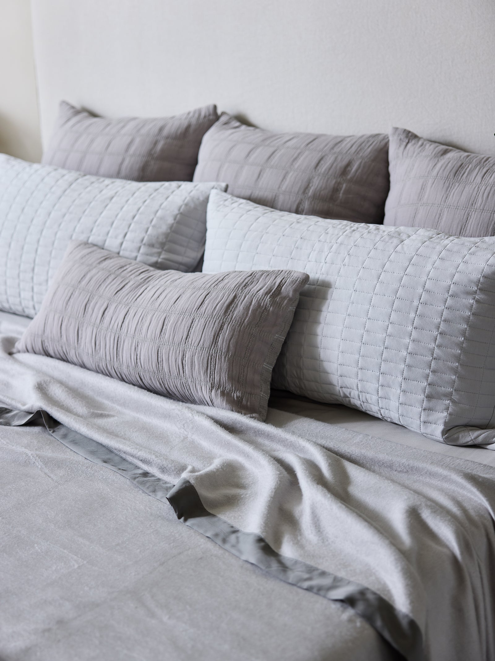 A neatly made bed with multiple light gray pillows of varying textures and patterns arranged against a white headboard. A soft gray comforter and a Cozy Earth Bamboo Blanket are draped across the bed, enhancing the monochromatic color scheme. 