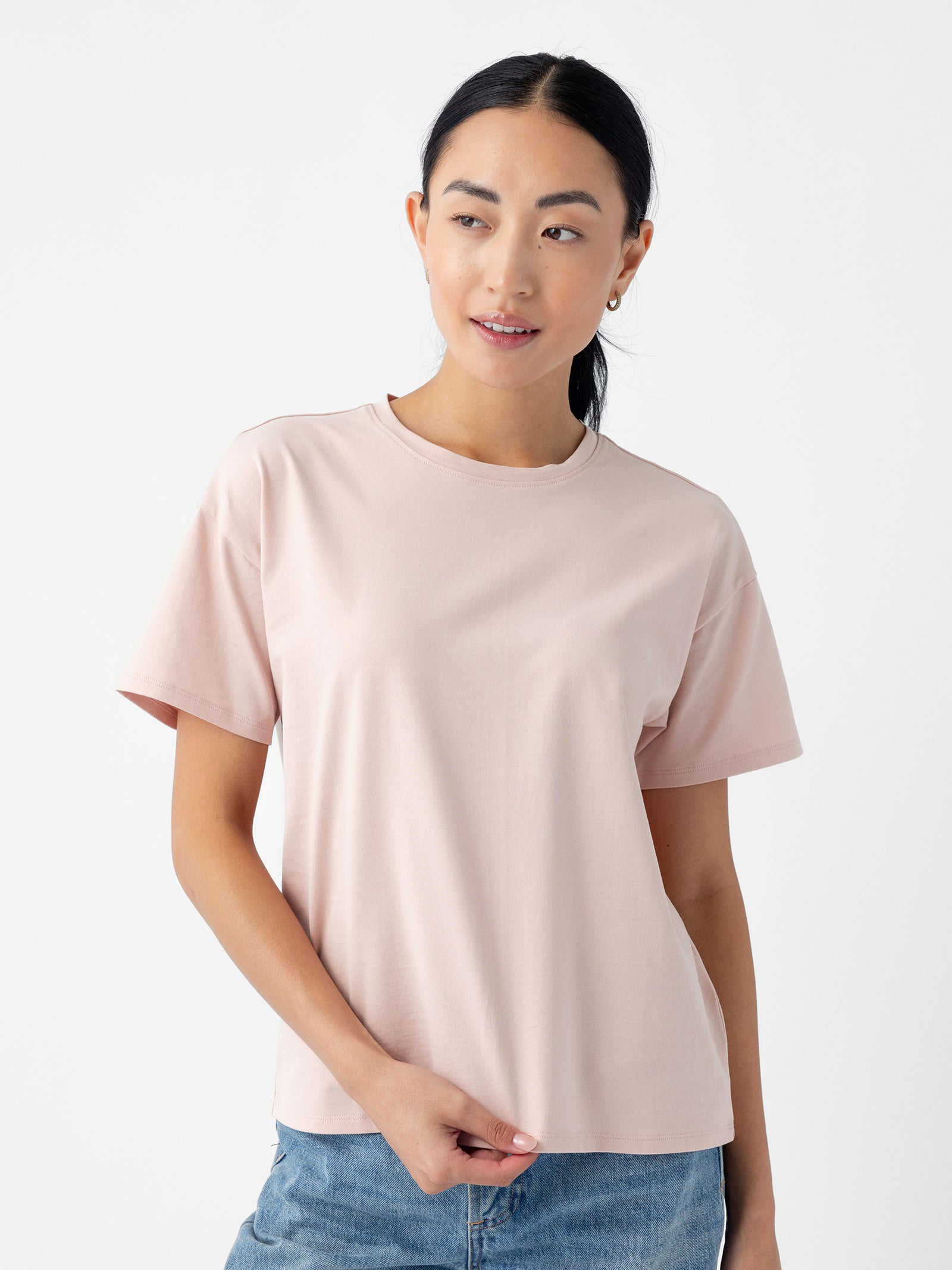 A person with long dark hair is wearing a light pink Women's All Day Tee from Cozy Earth and jeans. They are looking slightly to the side with a neutral expression, against a plain white background. 