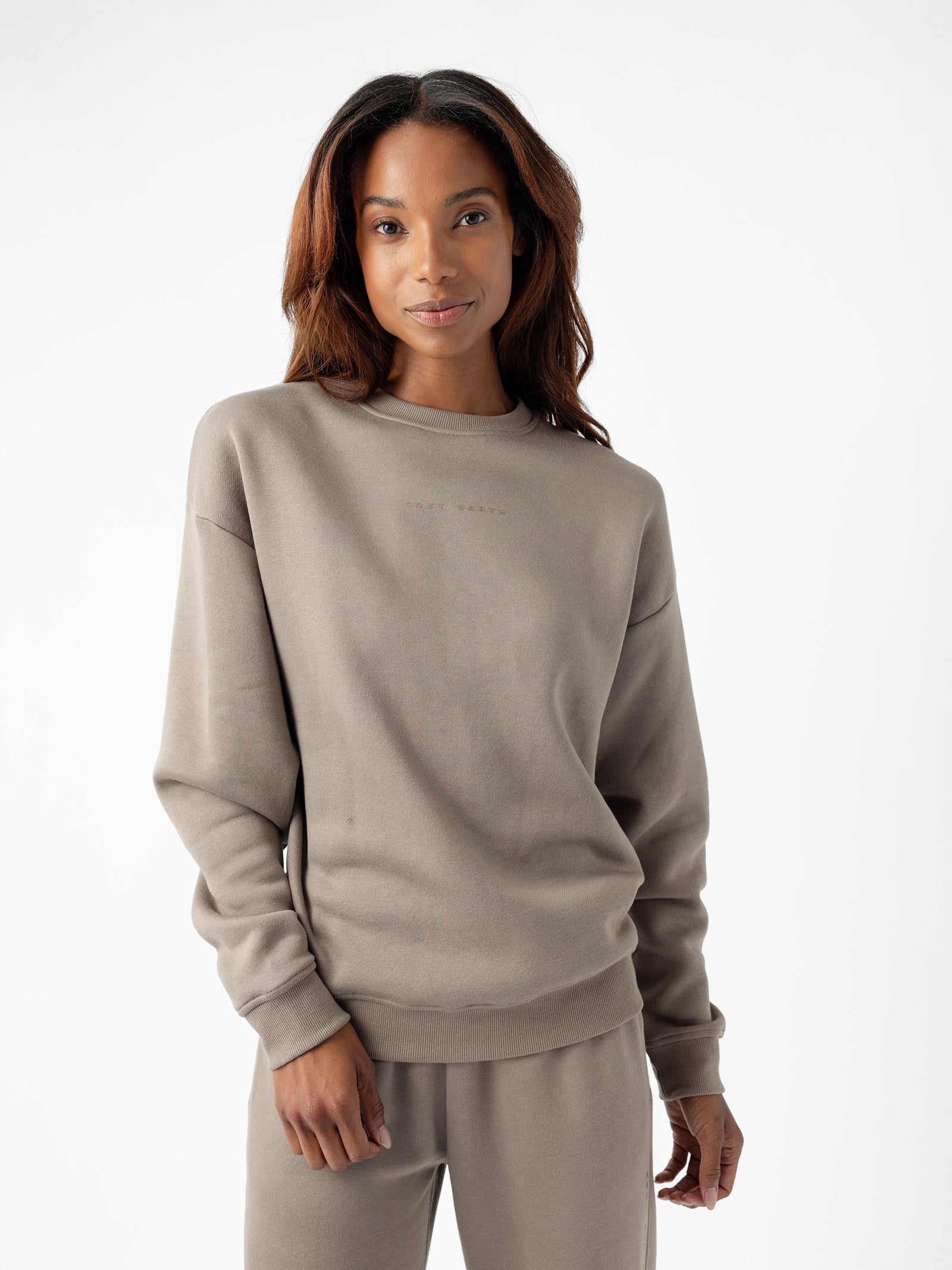 A person with long, wavy hair is wearing the Women's CityScape Crewneck and matching taupe-colored sweatpants from Cozy Earth. They are standing against a white background and smiling softly at the camera. The crewneck has subtle text on the chest. 