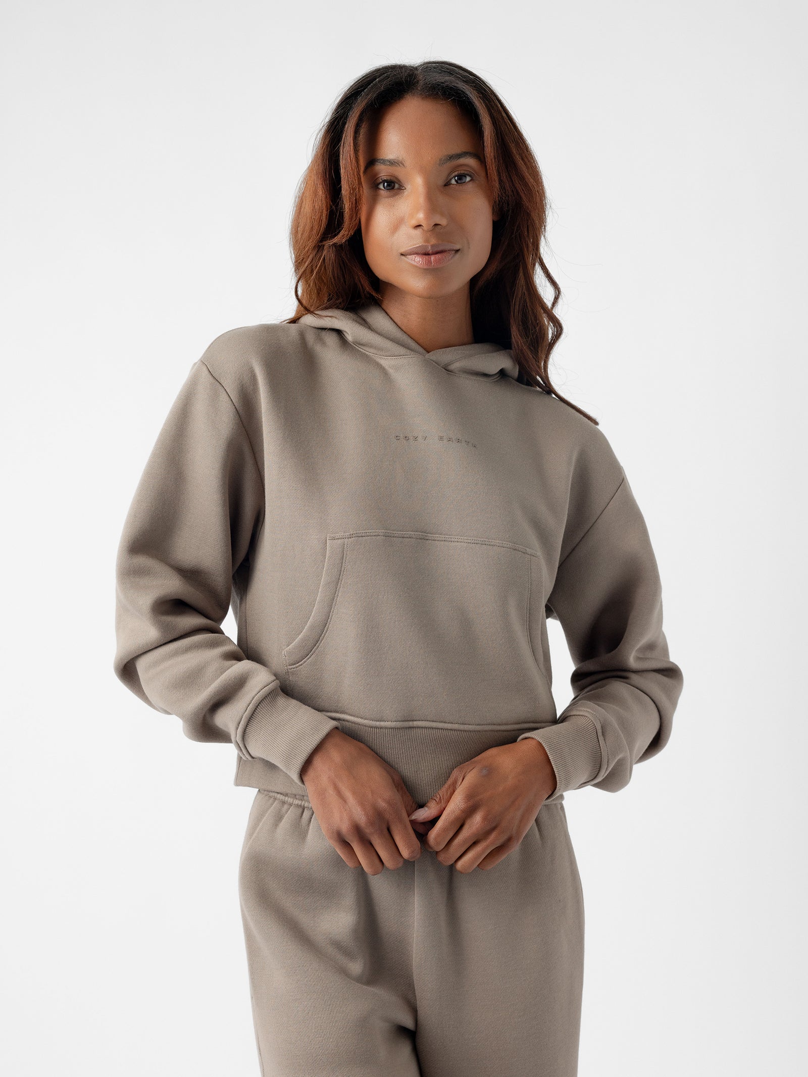 A person with long, wavy hair is wearing a light brown Women's CityScape Cropped Hoodie by Cozy Earth and matching pants. They have their hands in the front pocket of the hoodie and are posing in front of a plain, white background. 