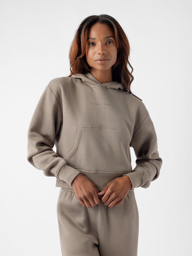 A person with long, wavy hair is wearing a light brown Women's CityScape Cropped Hoodie by Cozy Earth and matching pants. They have their hands in the front pocket of the hoodie and are posing in front of a plain, white background.