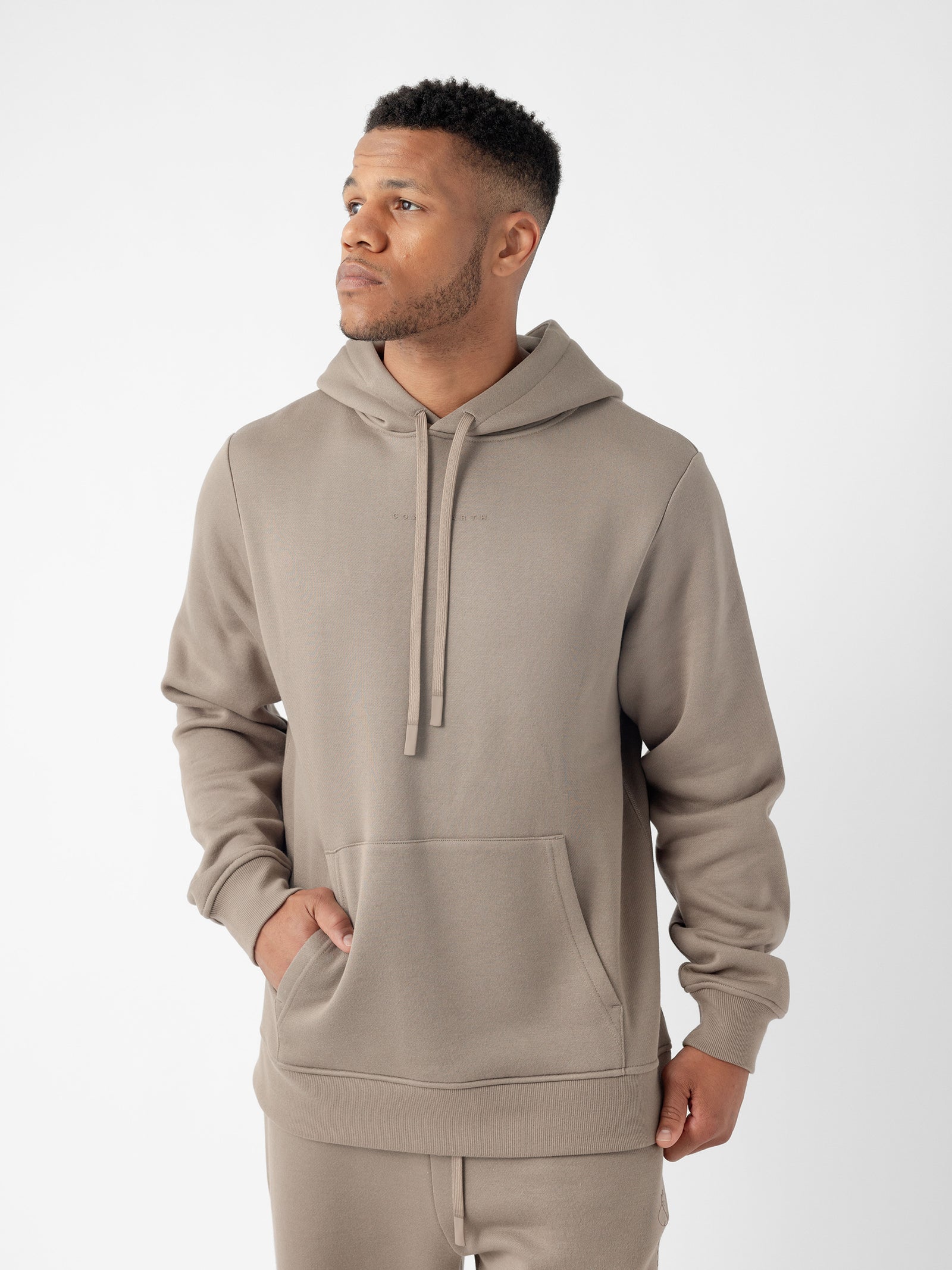 A man stands against a plain white background, wearing Cozy Earth's Men's CityScape Hoodie in a taupe color with a front pocket and matching joggers. He has short hair and is looking to his left with one hand partially in his pocket. 