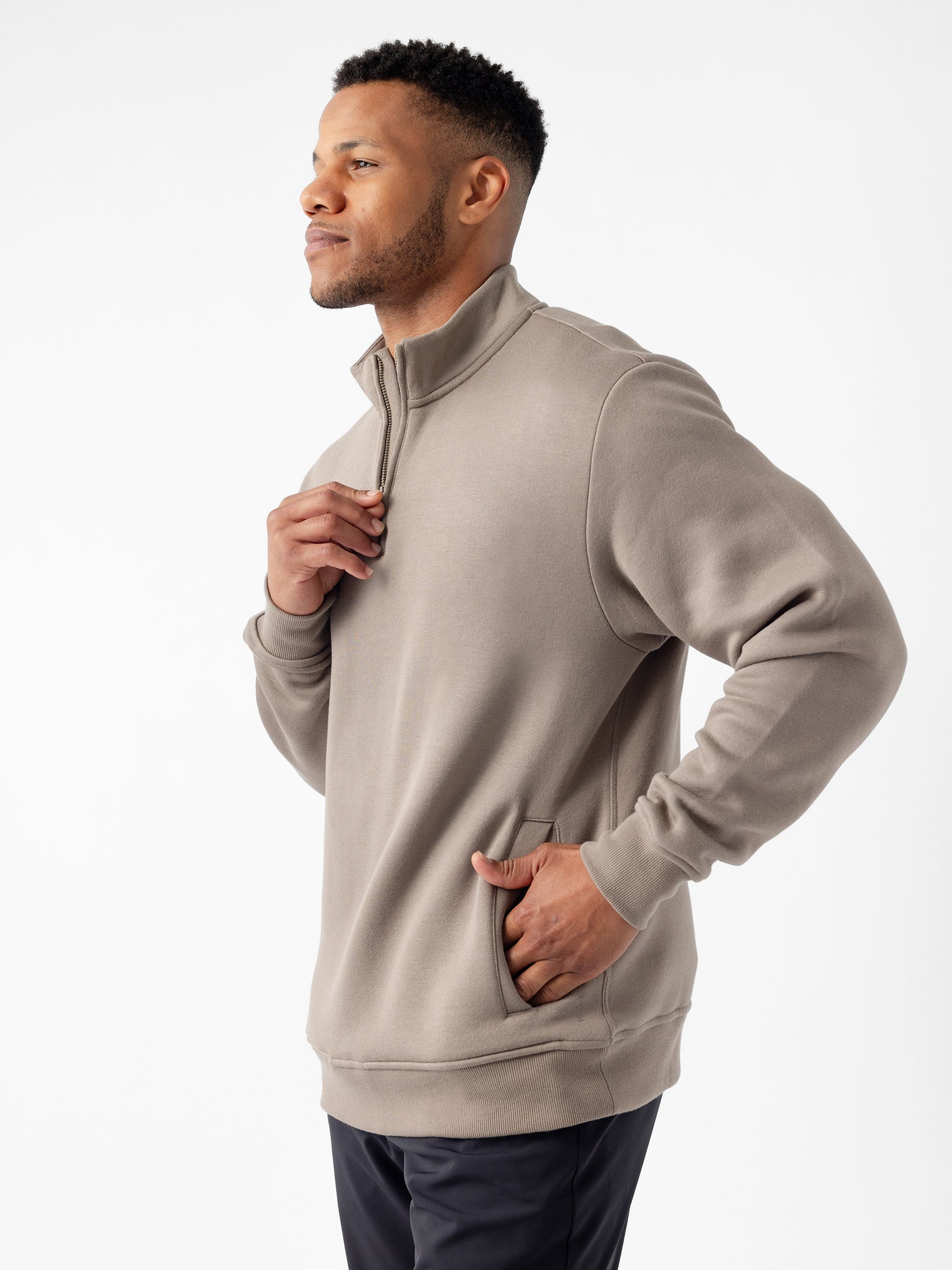 A person with short, dark hair and a trimmed beard is posing in a light gray Men's CityScape Quarter Zip by Cozy Earth. They are holding the collar with one hand and have the other hand in the pocket. They are looking confidently to the side against a plain white background. 