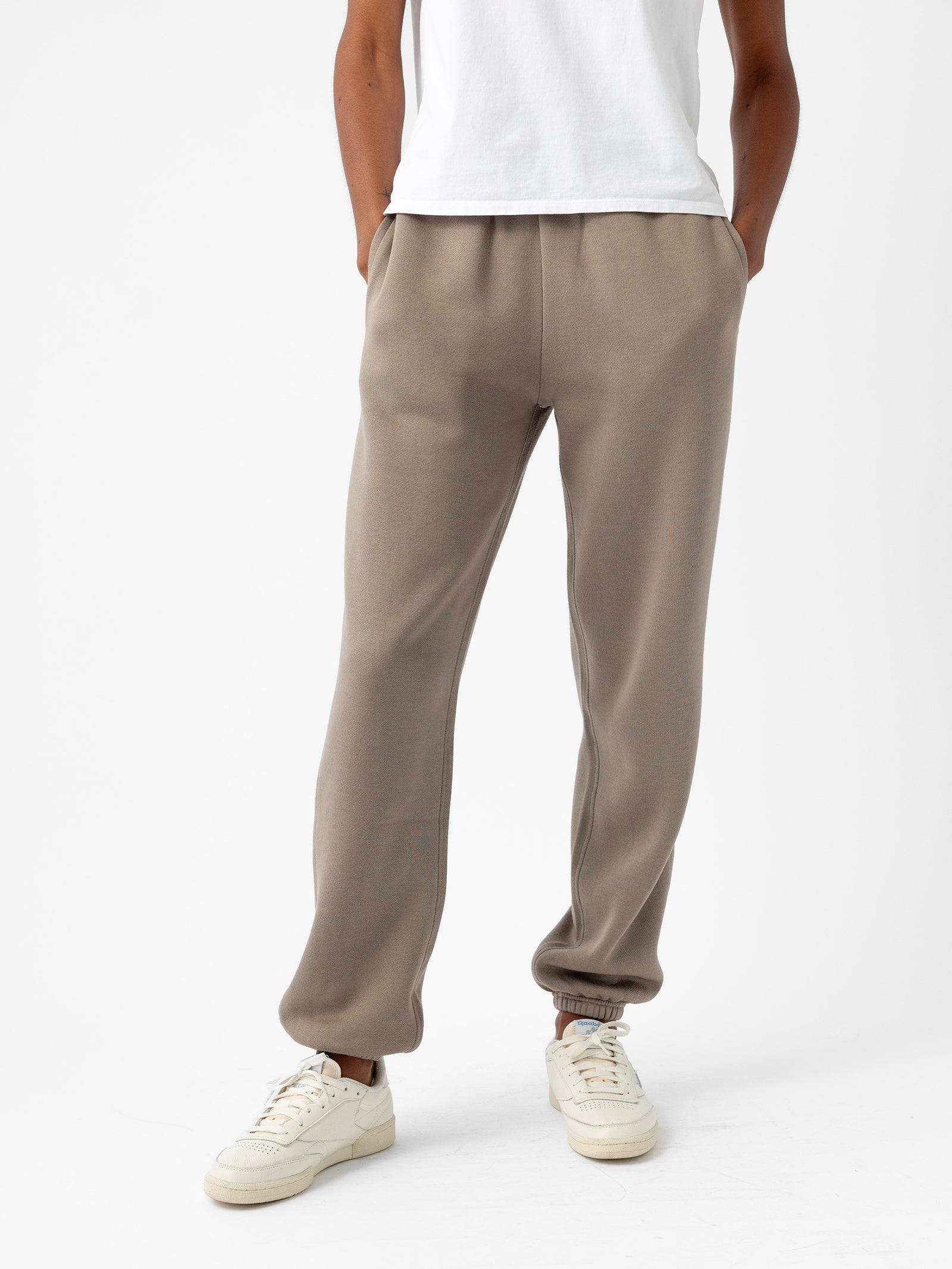 A person is wearing Women's CityScape Sweatpants in beige from Cozy Earth and a white t-shirt, with their hands in the pockets of the sweatpants. The person is also wearing white sneakers. The background is plain white. 