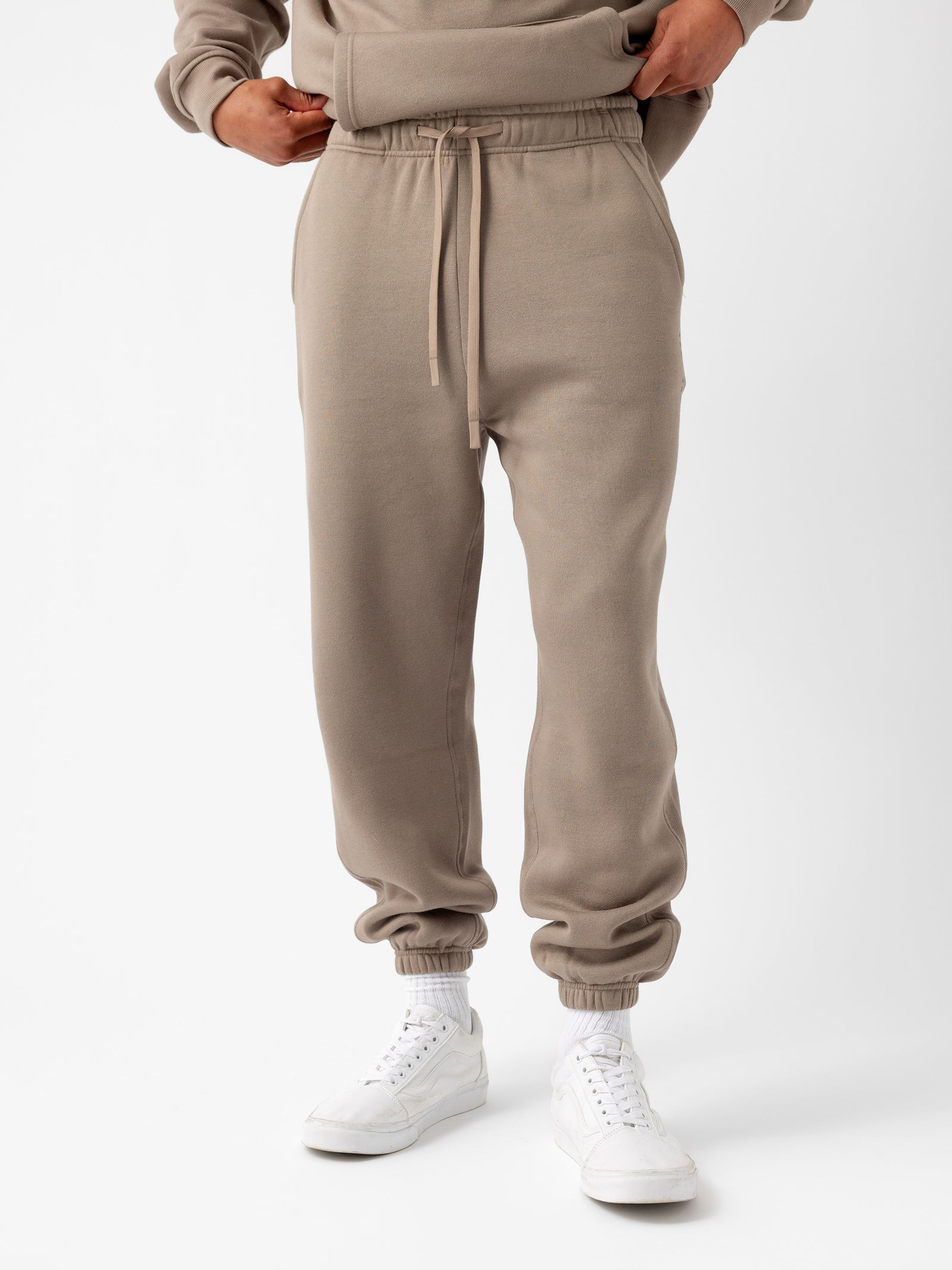 A person is wearing the Men's CityScape Sweatpant by Cozy Earth, which features a drawstring waist and cuffs at the ankles. They are also dressed in a matching beige sweatshirt and white sneakers paired with white socks. The background is plain white. 