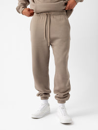 A person is wearing the Men's CityScape Sweatpant by Cozy Earth, which features a drawstring waist and cuffs at the ankles. They are also dressed in a matching beige sweatshirt and white sneakers paired with white socks. The background is plain white. |Color:Ebony