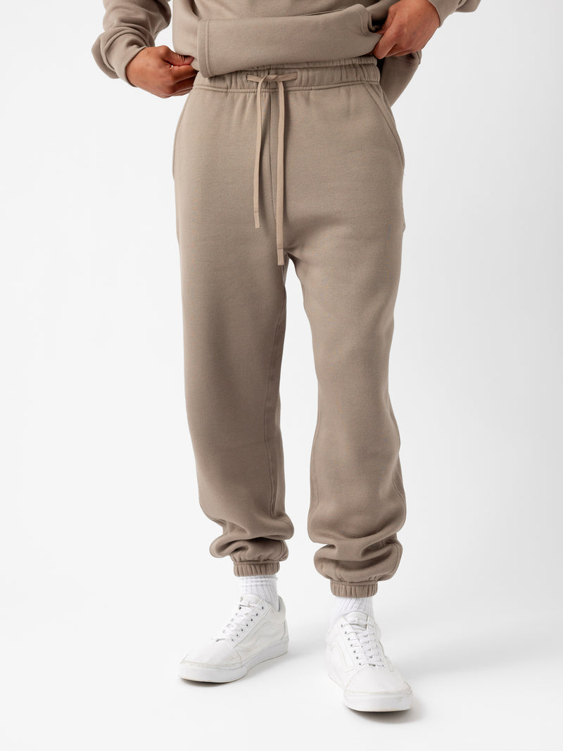 A person is wearing the Men's CityScape Sweatpant by Cozy Earth, which features a drawstring waist and cuffs at the ankles. They are also dressed in a matching beige sweatshirt and white sneakers paired with white socks. The background is plain white.