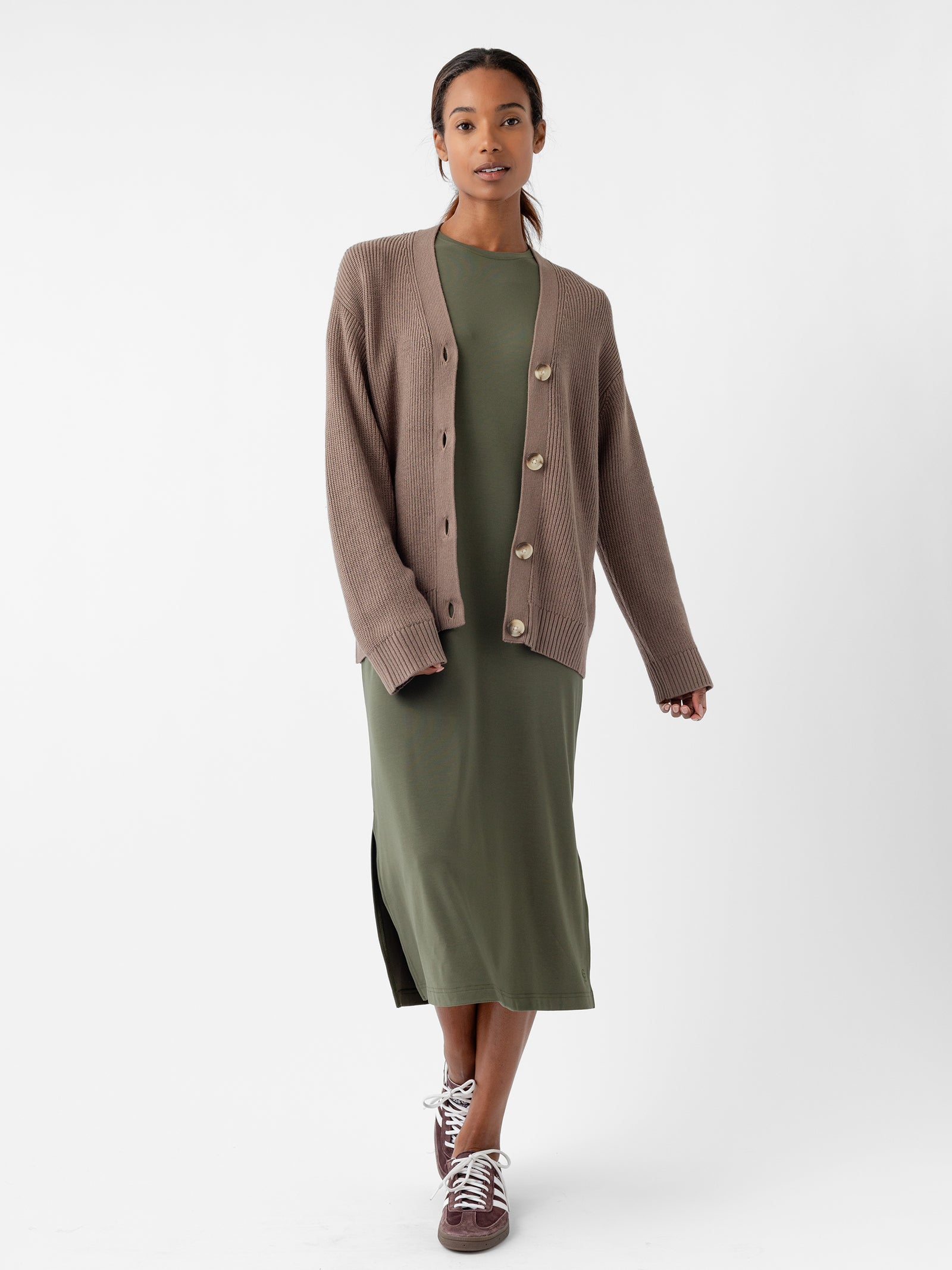 A person wearing Cozy Earth's Oversized Classic Cardigan in brown pairs it with a long, green dress while standing against a plain background. They have on maroon sneakers and maintain a neutral expression, presenting an outfit that is both casual and comfortable. 