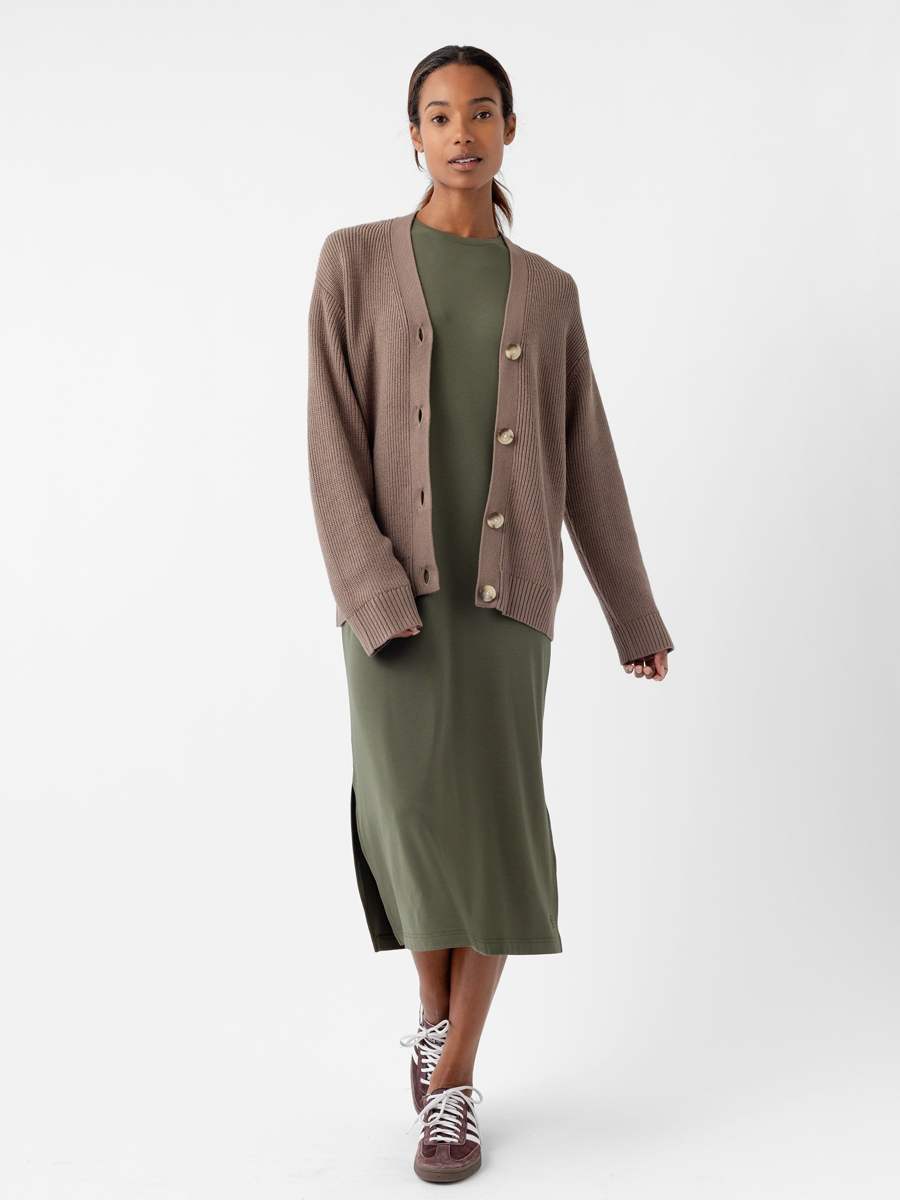 A person wearing Cozy Earth's Oversized Classic Cardigan in brown pairs it with a long, green dress while standing against a plain background. They have on maroon sneakers and maintain a neutral expression, presenting an outfit that is both casual and comfortable. |Color:Ebony