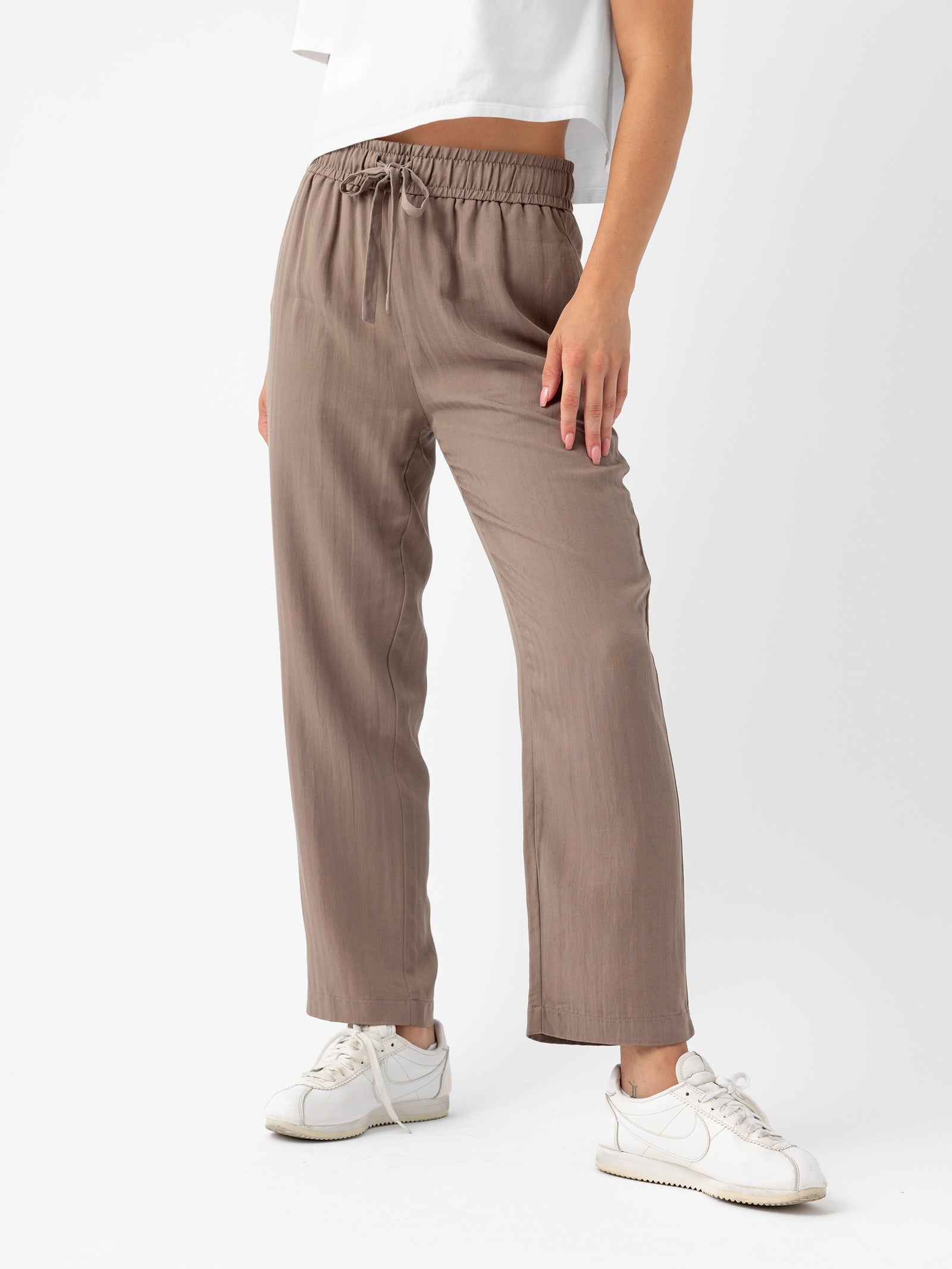 A person is standing against a white background wearing a white cropped top, the Women's Sunset Cropped Pant by Cozy Earth in beige, and white sneakers. The person’s right hand rests on their upper thigh, and their head is out of the frame. 