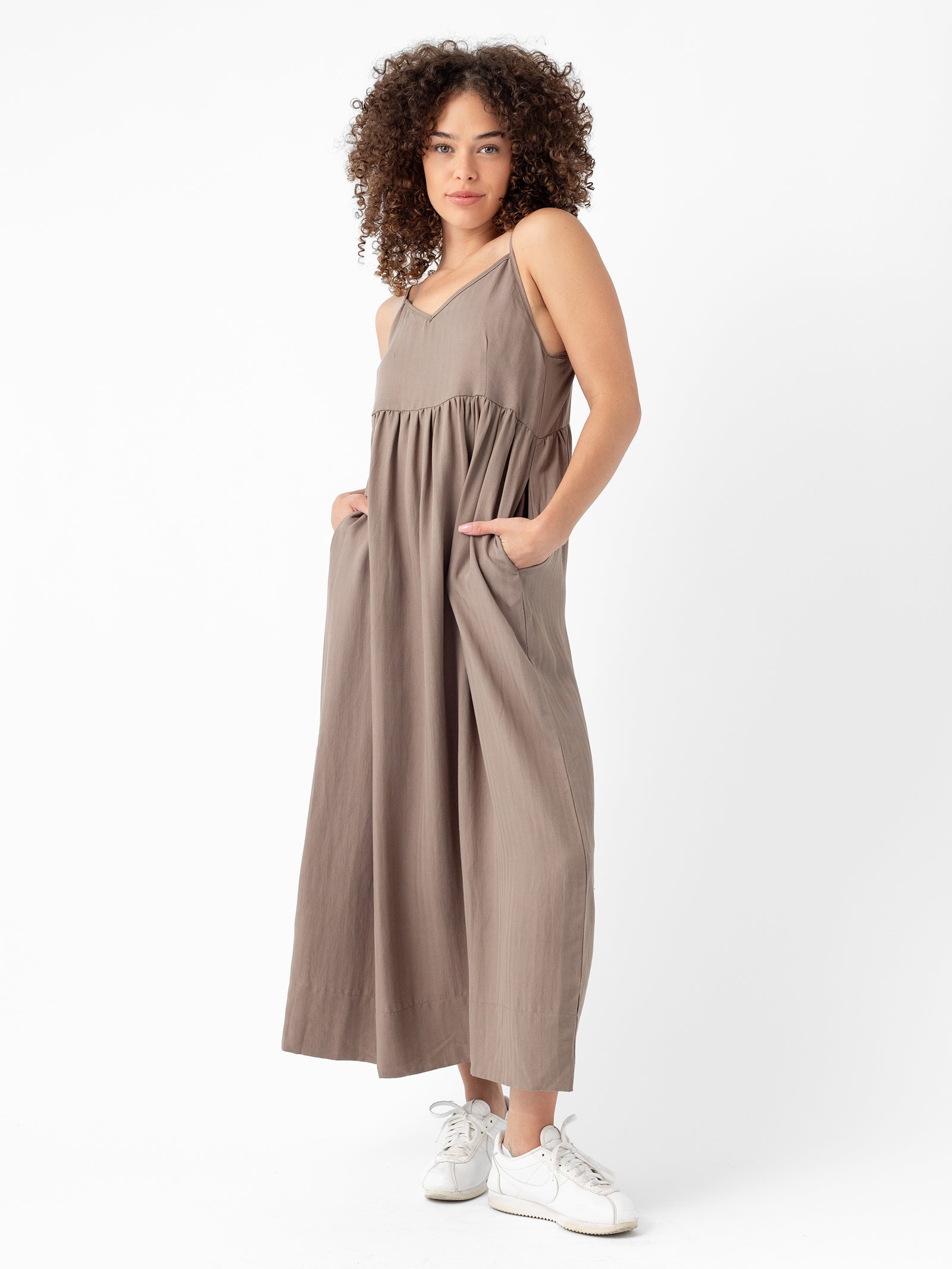 A person with curly hair wearing a Cozy Earth Women's Sunset Gathered Dress, which is loose, sleeveless, ankle-length and in a light brown shade, stands with hands in the dress pockets. They are wearing white sneakers and posing against a plain white background. 