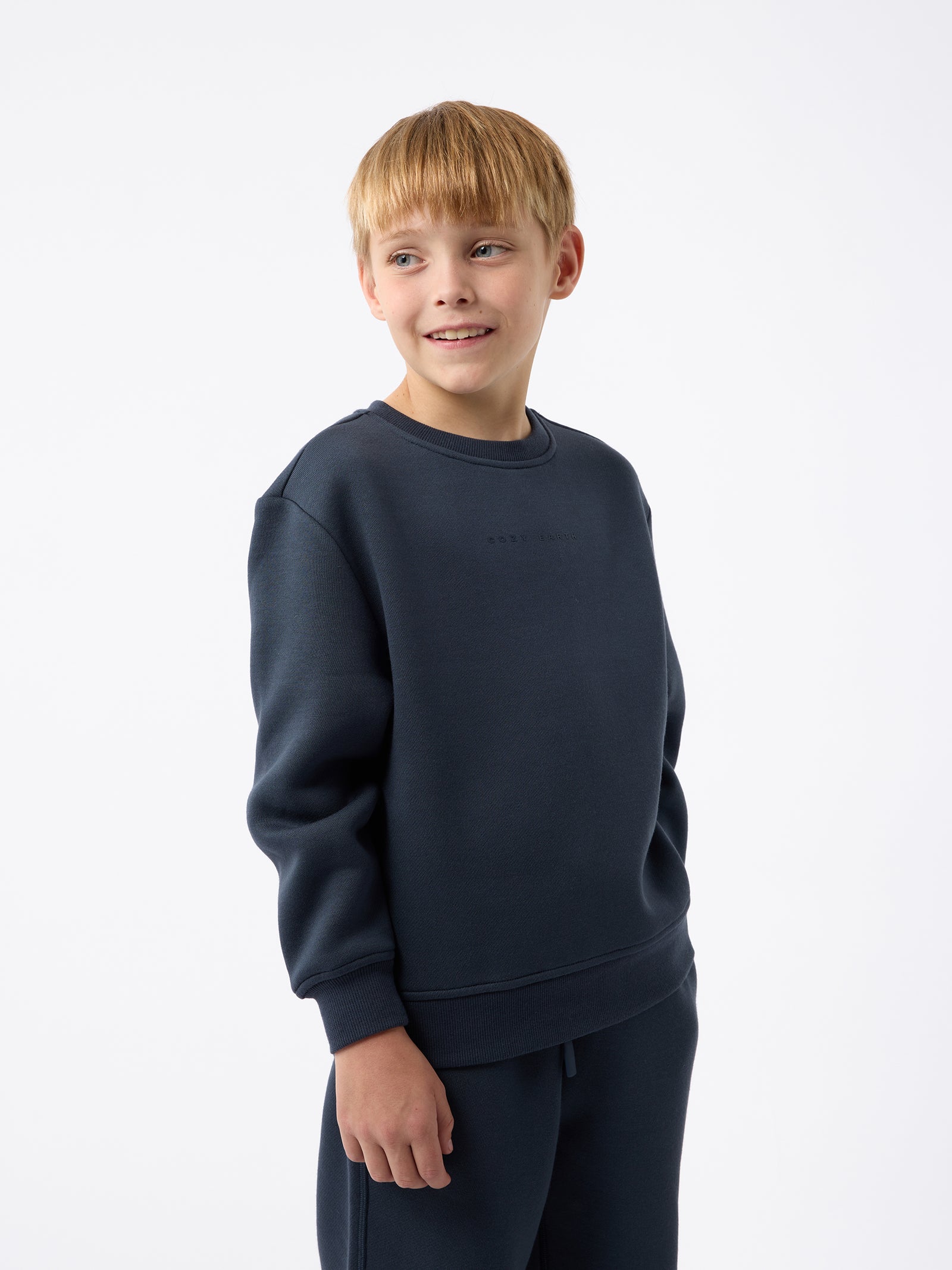 A young boy with blond hair is wearing the Cozy Earth Kid's CityScape Crewneck sweatshirt along with matching pants. He is standing, smiling, and looking slightly to the side against a plain white background. 