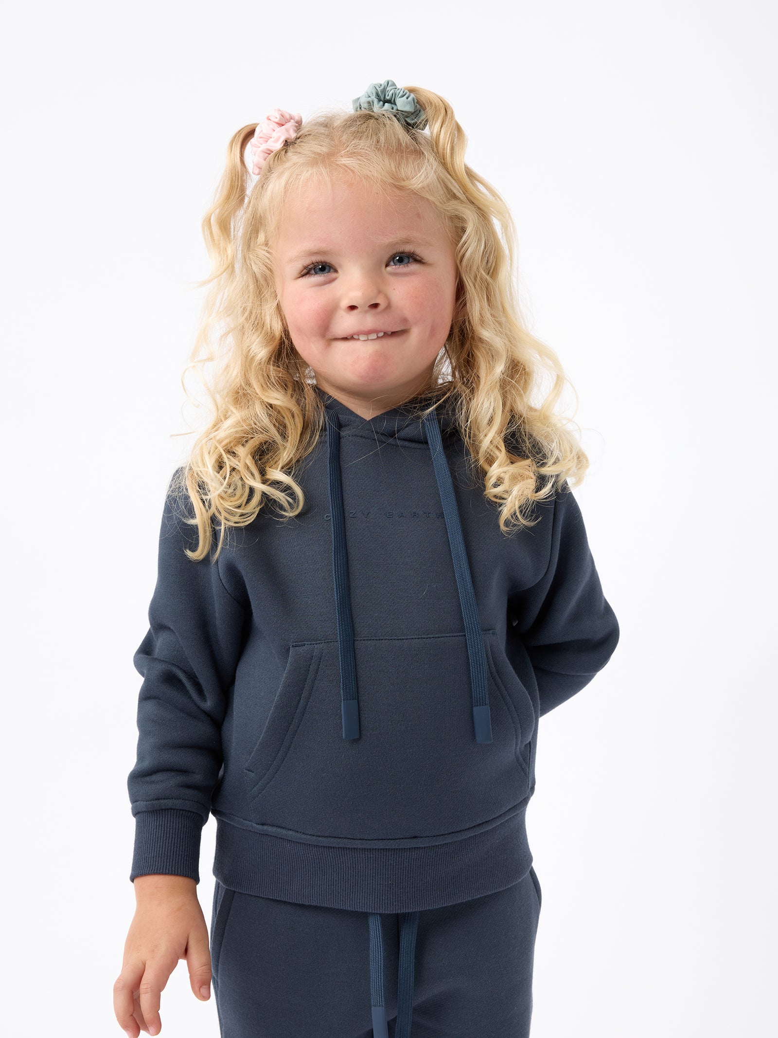 A young child with long blonde hair smiles while wearing a navy blue Kid's CityScape Hoodie by Cozy Earth and matching pants. She has two hair ties, one pink and one blue. The background is plain white. 