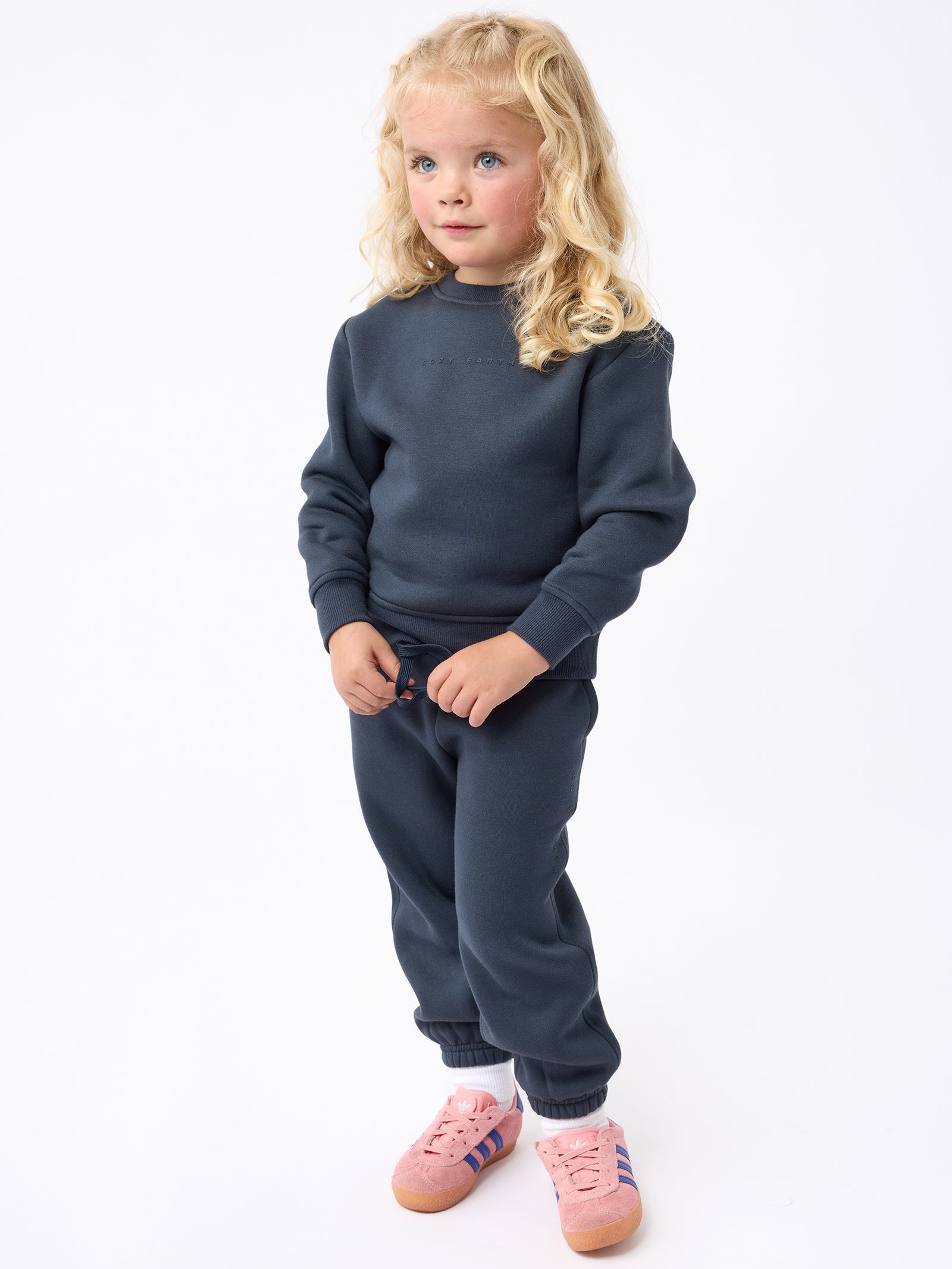 A young child with long blonde hair stands against a white background, dressed in a dark blue tracksuit and Cozy Earth's Kid's CityScape Sweatpant. They are wearing pink sneakers with purple stripes and have a neutral expression, with their hands tucked into the pockets of the pants. 